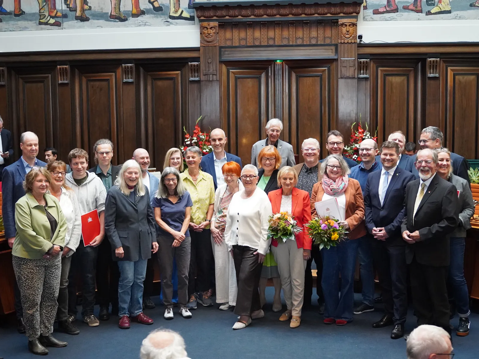 Eine Gruppe von Menschen stehen in einem Raum frontal zur Kamera, einige halten Urkunden oder Blumen in den H&auml;nden.