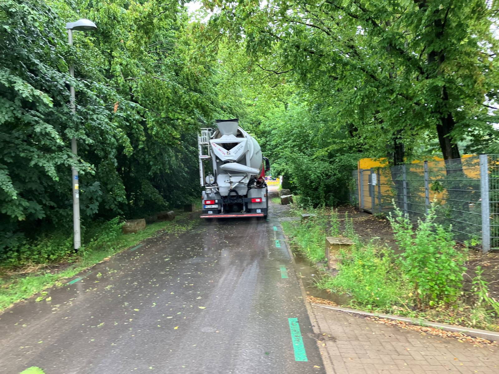 Auf einem asphaltierten Weg ist von hinten ein fahrendes Betonmisch-Fahrzeug zu sehen, rechts grüne Bäume und Büsche mit einer Laterne, rechts Bäume und ein Zaun, vorn eine Einfahrt.