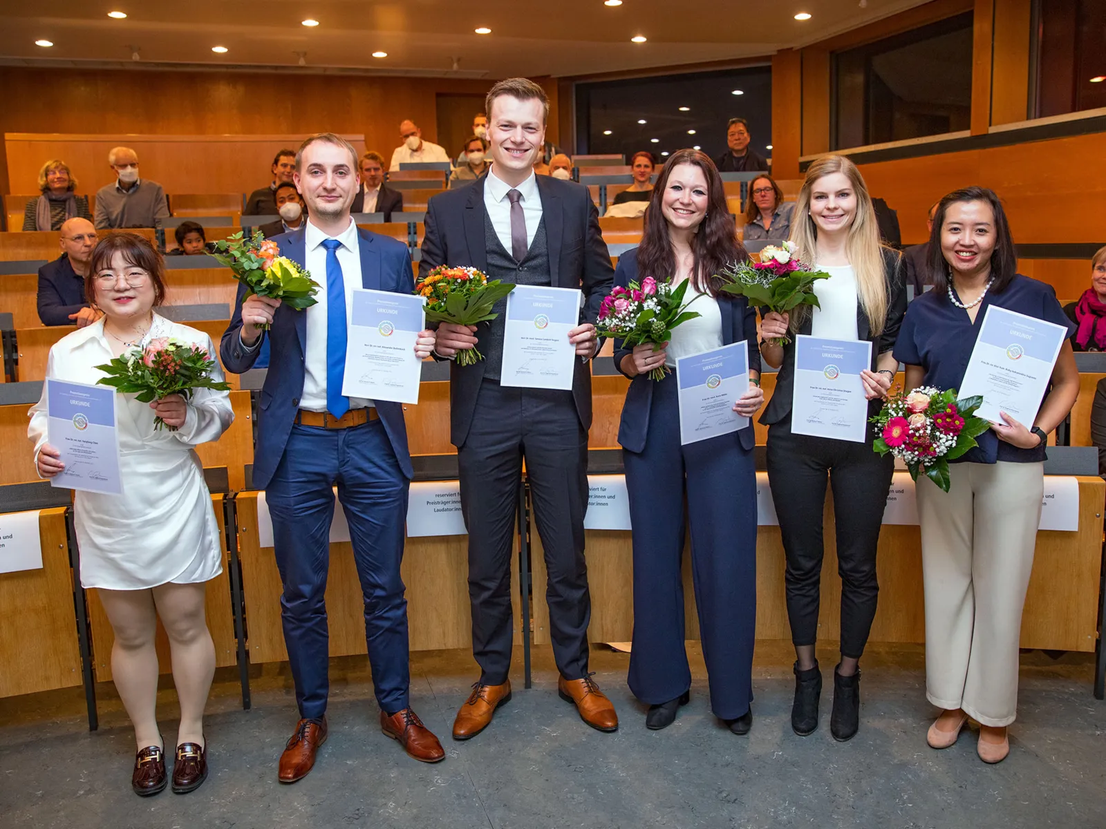 Vier junge Frauen und Männer mit Blumen und Urkunden in der Hand.
