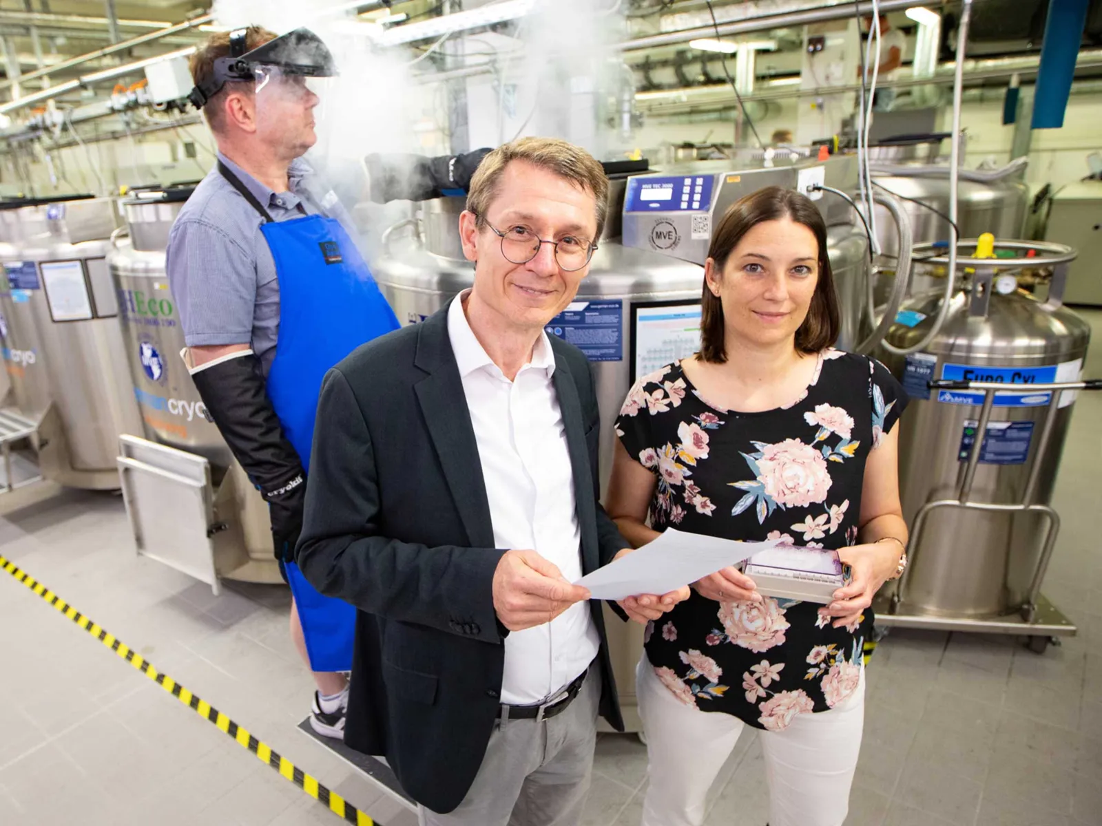 Ein Mann und eine Frau, im Hintergrund ein weiterer Mann mit Schutzkleidung.