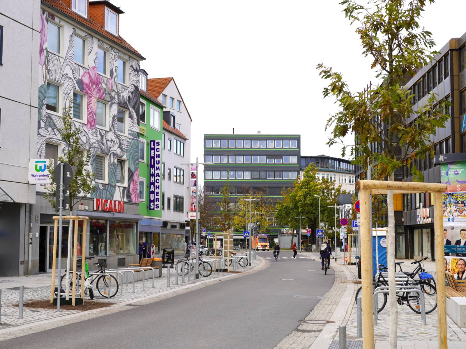 Eine Straße mit zwei Radfahrenden, rechts und links Fahrradbügel, kleine mit Holz geschützte Bäume und Gebäude.