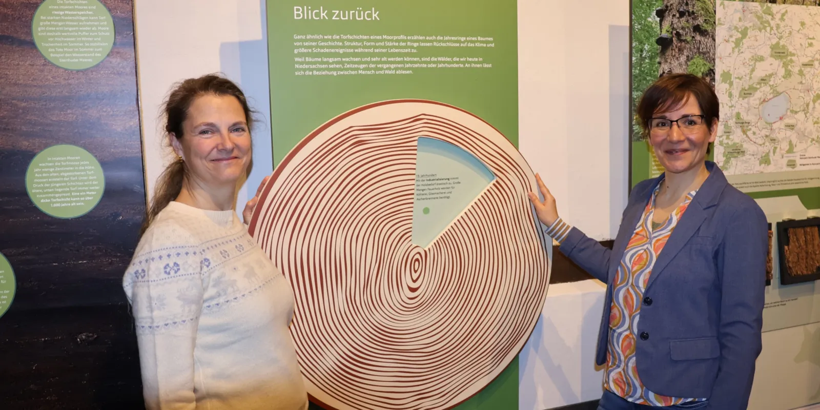 Zwei Frauen stehen in einem Ausstellungsbereich mit Mitmach-Aktionen an einer Drehscheibe in Baumscheibenoptik mit Jahresringen. Ein Sichtfenster in der Drehscheibe l&auml;sst immer nur einen kleinen Ausschnitt des darunter liegenden Textbereiches frei. An der Wand dar&uuml;ber steht unter anderem: "Blick zur&uuml;ck"