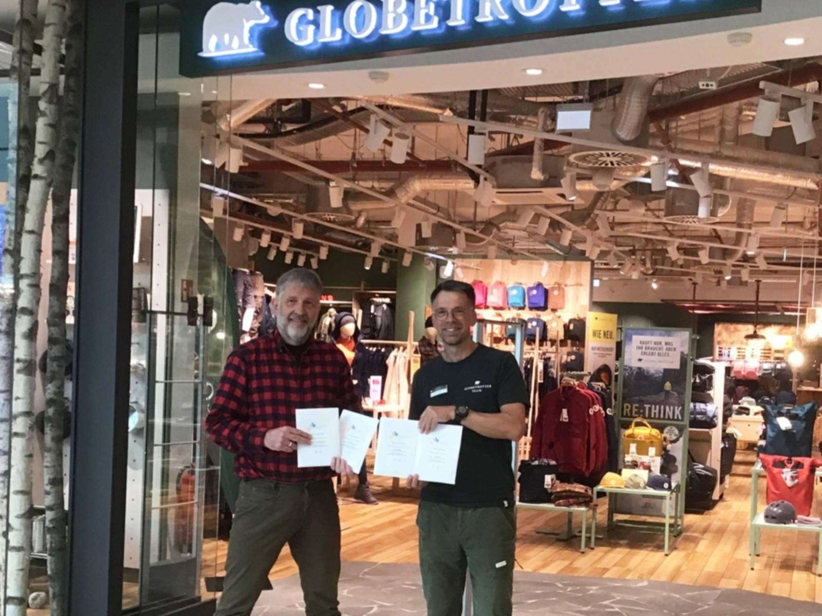 Zwei Männer vor einem Ladeneingang präsentieren vier Papiertüten, die sich vor sich halten.