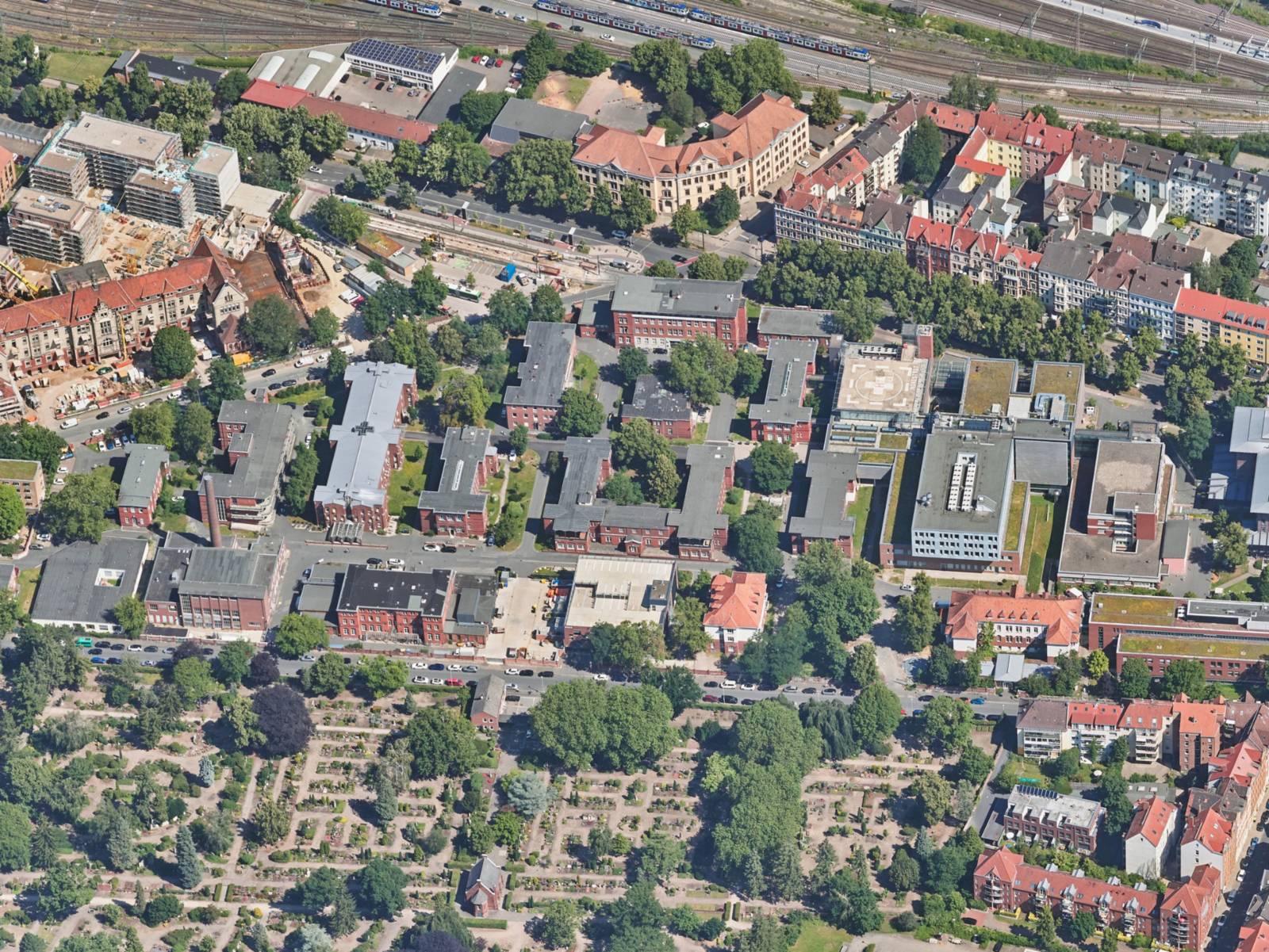Luftbild auf den Bereich einer Stadt mit mehreren Geb&auml;uden, Wegen und Stra&szlig;en, B&auml;umen. Vorne Gr&uuml;nfl&auml;che in grafischen Rechtecken, hinten Bahnschienen.