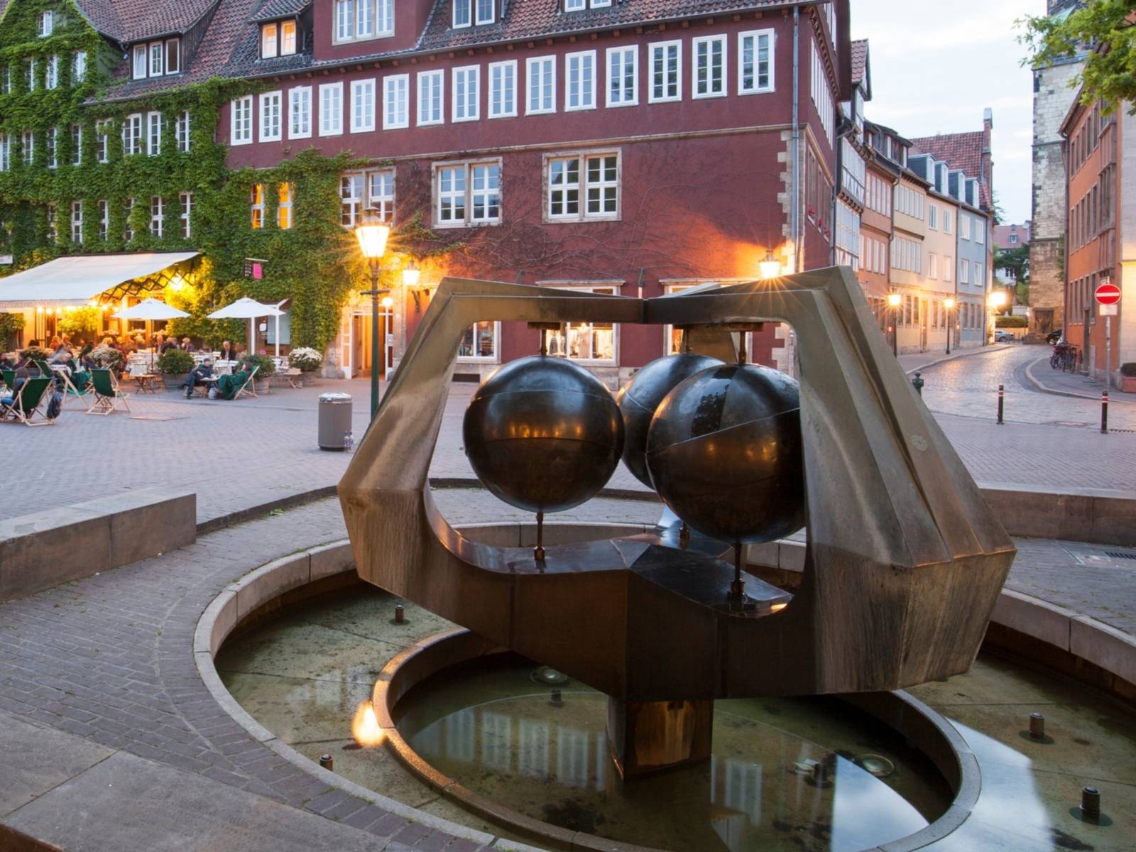 Brunnnen mit einer Installation aus drei Kugeln, im Hintergrund sitzen Leute auf Liegestühlen auf einem von historischen Gebäuden gesäumten Platz.