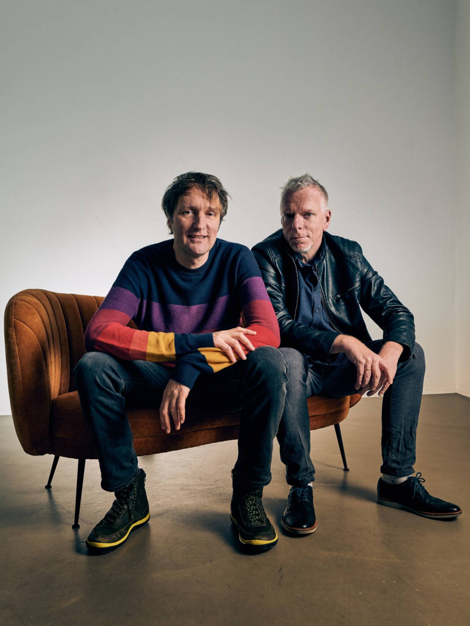 Zwei Männer sitzen auf einem Polstermöbelstück und schauen in die Kamera.
