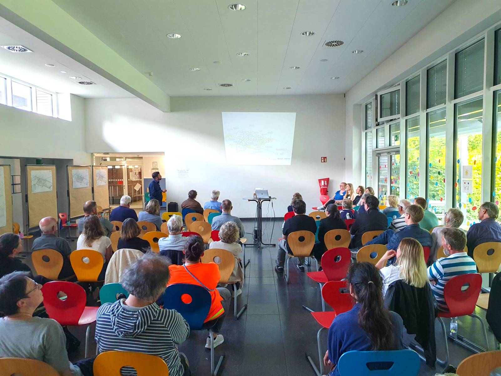 Menschen sitzen in Stuhlreihen in einem Raum und schauen auf eine beleuchtete Wand mit einer Pr&auml;sentation, daneben steht ein Mann.