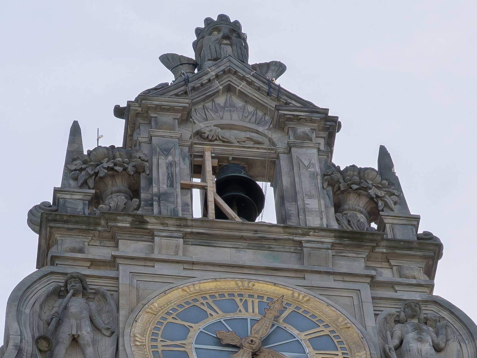 Detailaufnahme von der Spitze eines Bauwerks mit Zierelementen wie einem Kopf, Blumenk&uuml;beln und zwei Engeln mit Musikinstrumenten um eine Ohr mit goldenem Zifferblatt. Dar&uuml;ber eine Glocke in einem freien Fenster, das eine Holzkonstruktion abst&uuml;tzt.