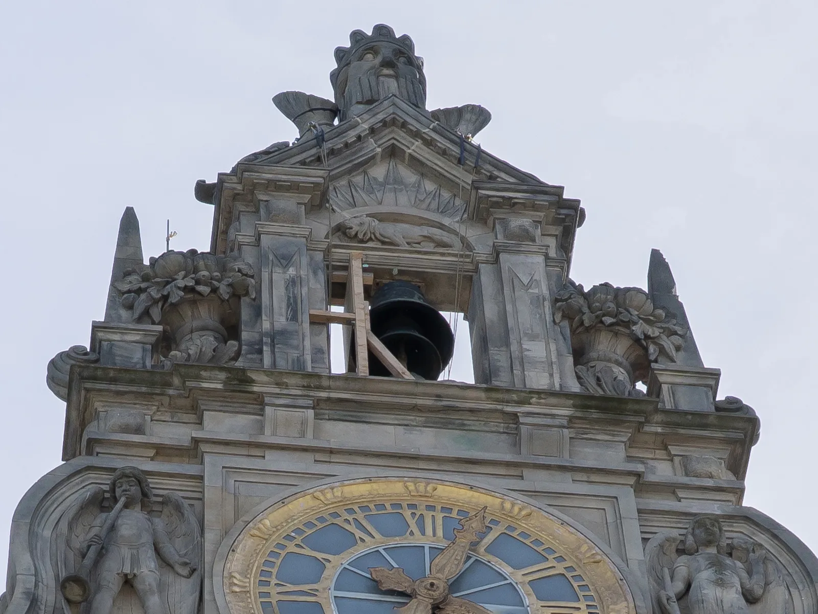 Detailaufnahme von der Spitze eines Bauwerks mit Zierelementen wie einem Kopf, Blumenk&uuml;beln und zwei Engeln mit Musikinstrumenten um eine Ohr mit goldenem Zifferblatt. Dar&uuml;ber eine Glocke in einem freien Fenster, das eine Holzkonstruktion abst&uuml;tzt.