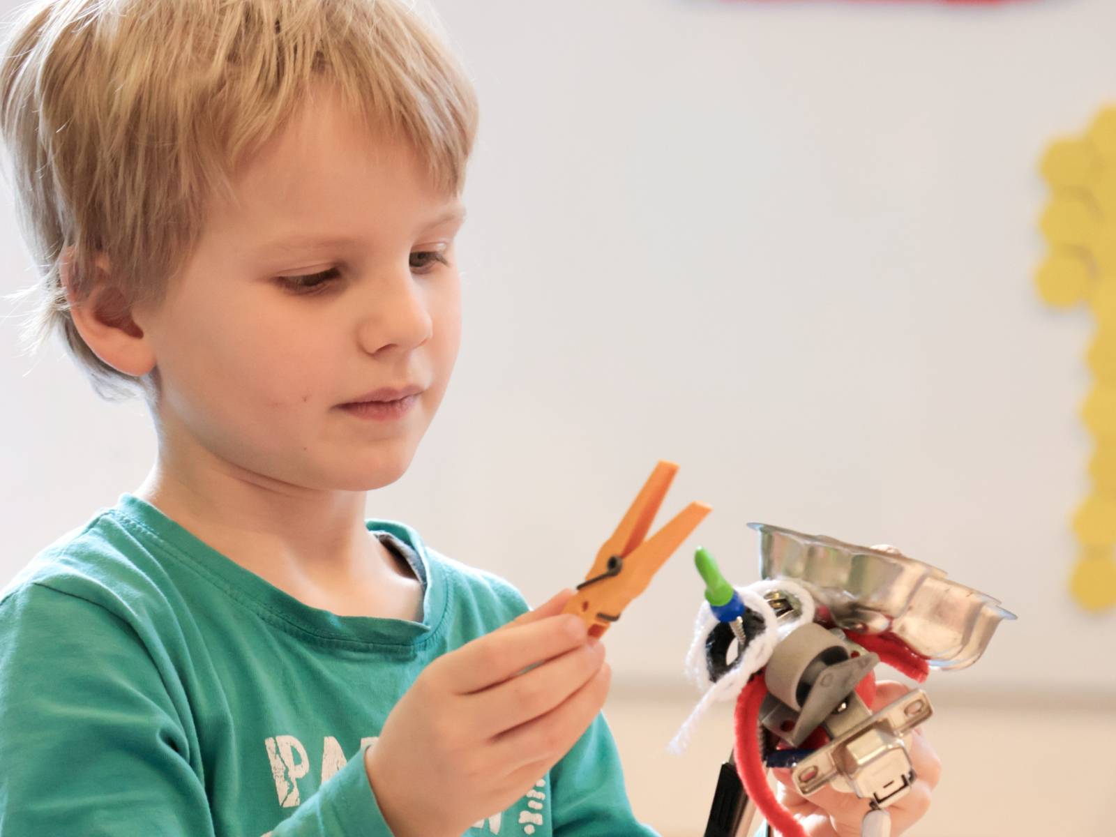 Ein Junge hält in der linken Hand einen Magneten. An diesem Magneten halten bereits verschiedene Gegenstände, zum Beispiel ein Spielzeug mit Metallfeder, eine Rolle für Möbel, ein Pfeifenreiniger und ein Möbelscharnier. In der rechten Hand hält der Junge eine Wäscheklammer.