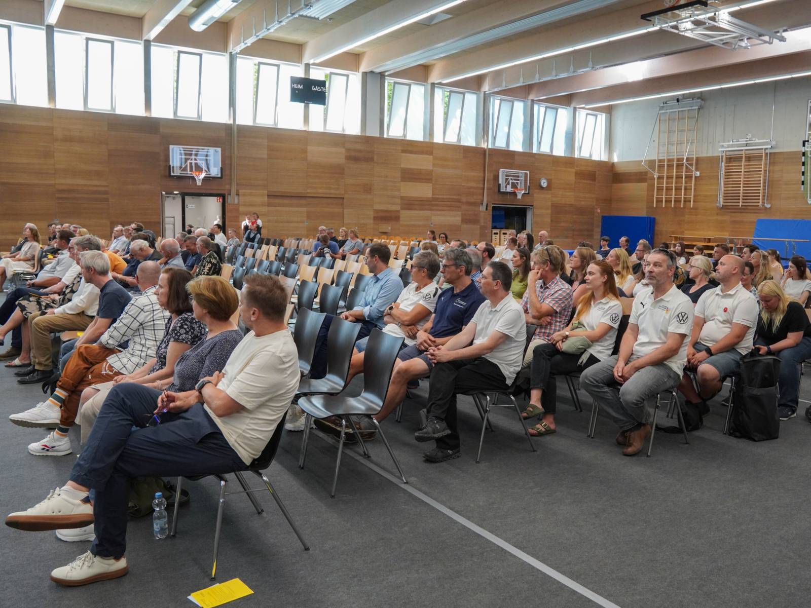 Menschen sitzen in Stuhlreihen in einer Halle mit Fenstern im oberen Bereich.