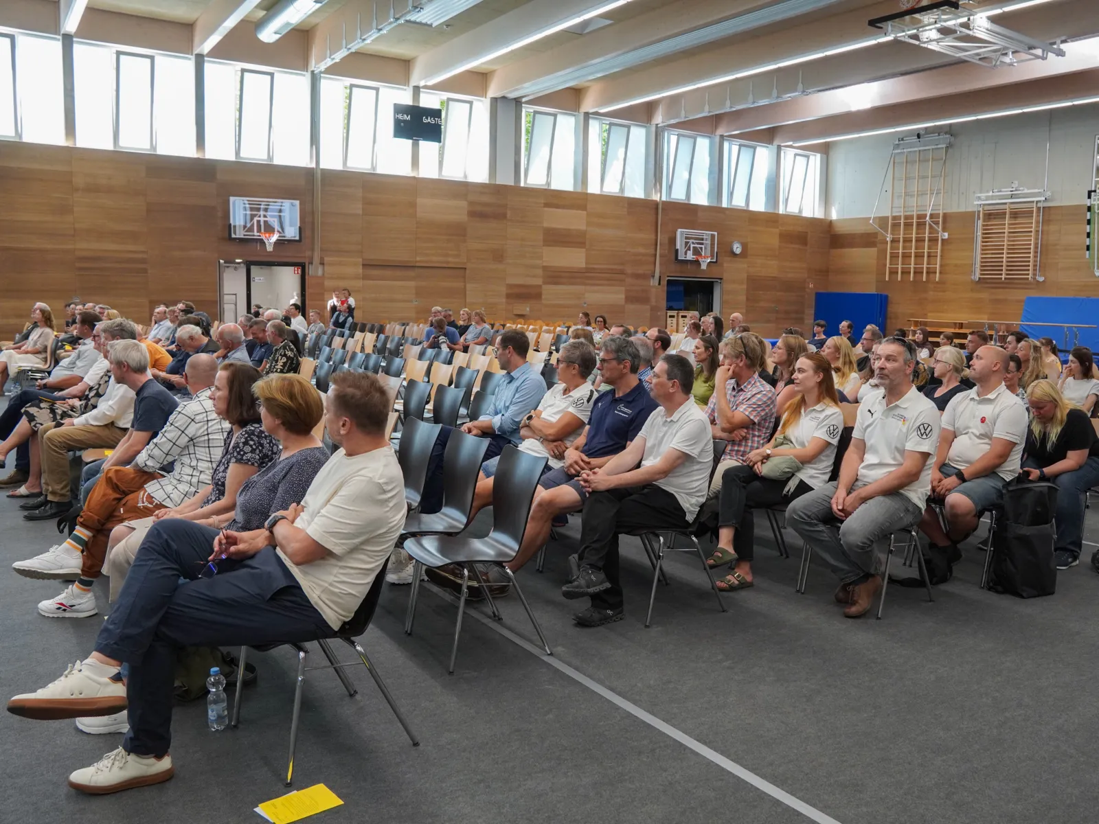 Menschen sitzen in Stuhlreihen in einer Halle mit Fenstern im oberen Bereich.