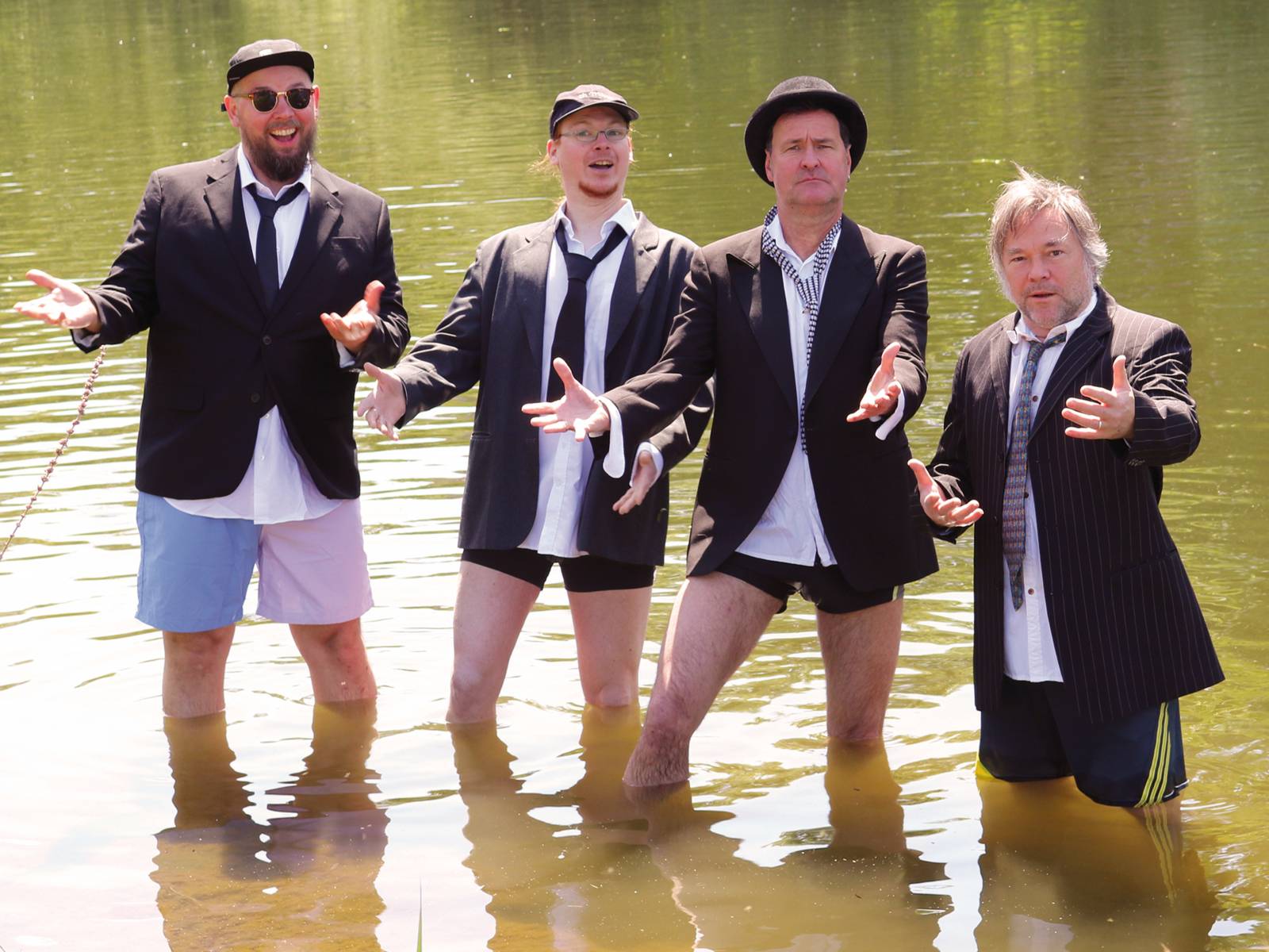 Vier Männer tragen kombinieren Badehosen und Sakkos mit Krawatten und weißen Hemden und singen in einem See.