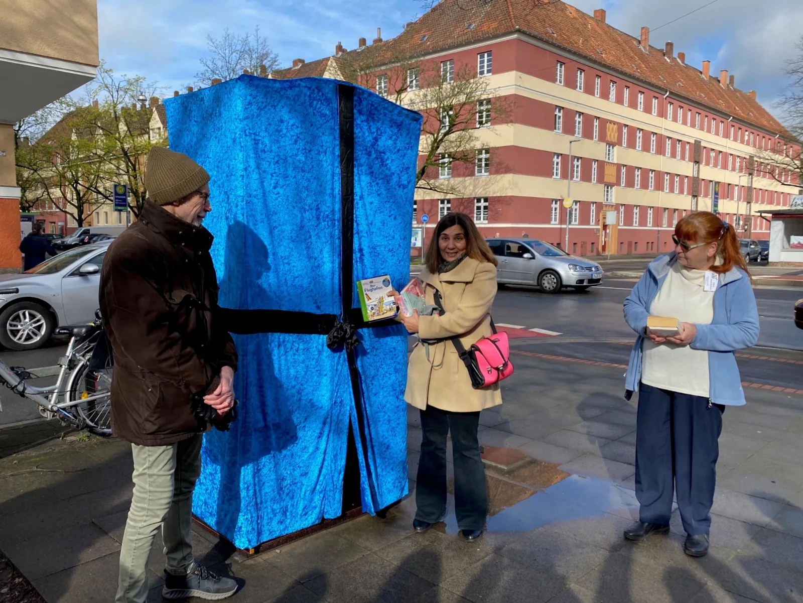 Auf dem Bild sind links ein Mann, mitte eine Frau und rechts eine Frau auf einem Gehweg zu sehen. Hinter ihnen steht ein blau verhüllter Bücherschrank.