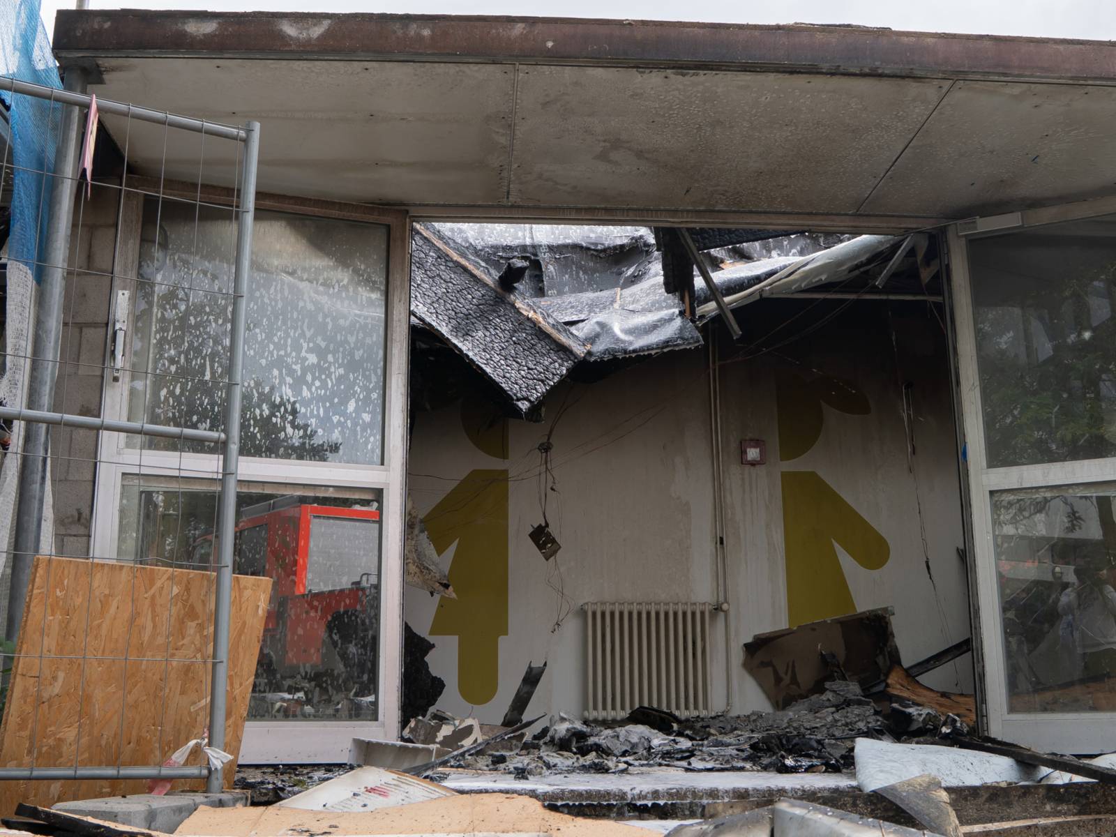 Ein offener Gebäudeteil mit Blick in einen Flur und aufgezeichneten Mädchen- und Jungen-Symbolen, darüber und an den Seiten herabgefallene, verrußte Decken, Türen und Gebäudeteile, unten herabgefallene Teile und weißer Löschschaum.