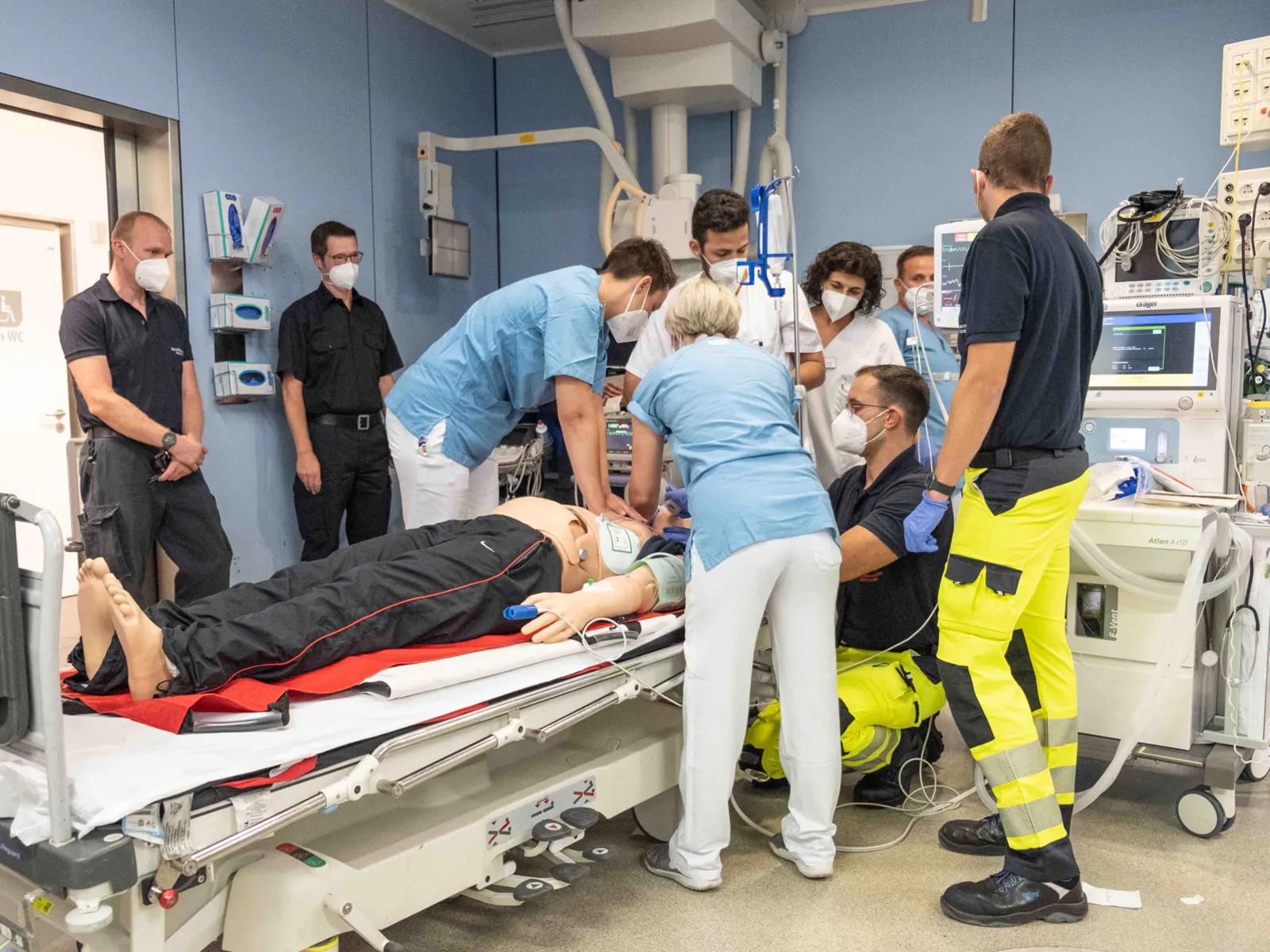 Verschiedene Personen befinden sich in einer Notaufnahme um eine Liege, auf der eine menschengroße Puppe liegt.