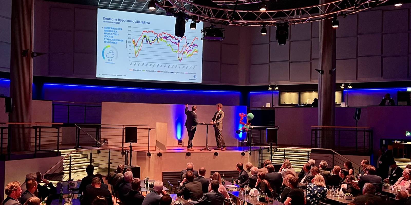 Zwei Personen pr&auml;sentieren bei einer Veranstaltung vor Publikum aktuelle Zahlen, Daten und Fakten.