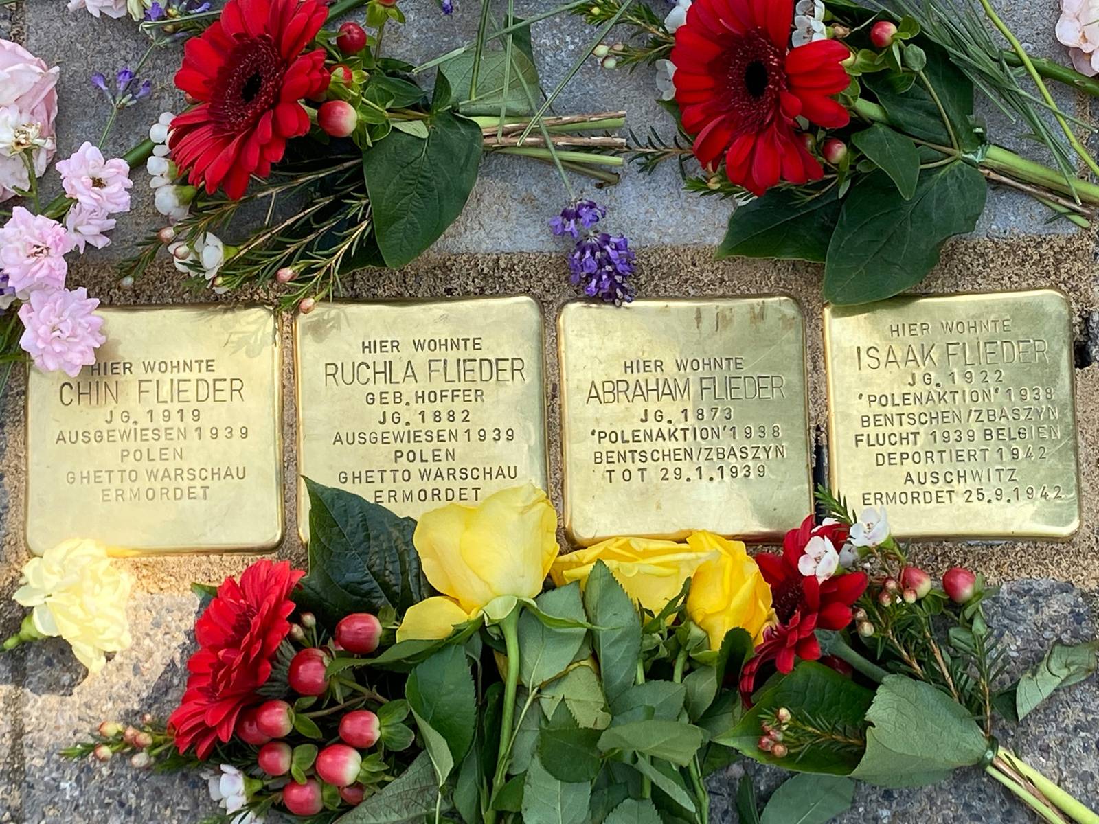 Vier goldene, quadratische Steine mit Inschrift im Boden: Chin Flieder, Ruchla Flieder, Abraham Flieder und Isaak Flieder.  Blumen drum herum.