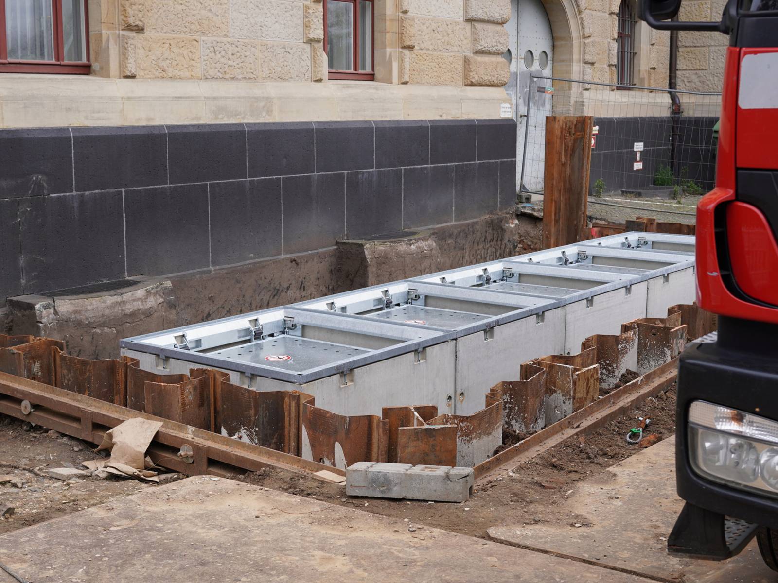 Vor einer Hauswand sind Betonbehälter in eine Baugrube eingelassen. Rechts ist der Scheinwerfer und ein Teil eines LKW zu erkennen.
