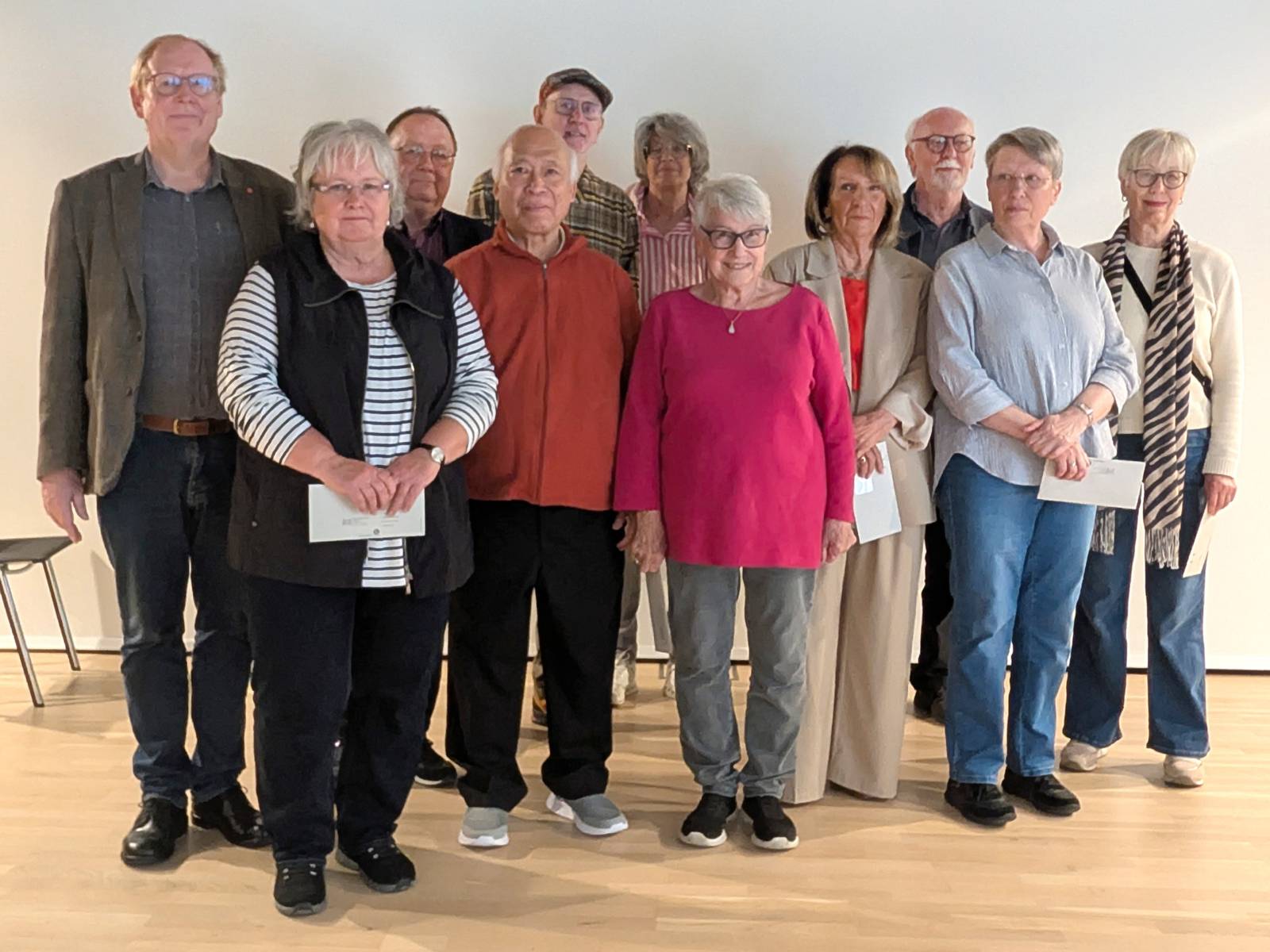 Eine Gruppe aus 13 Personen steht in einem Raum auf einem Holzboden vor einem weißen Banner. Die Frauen und Männer sind über 60 Jahre alt.