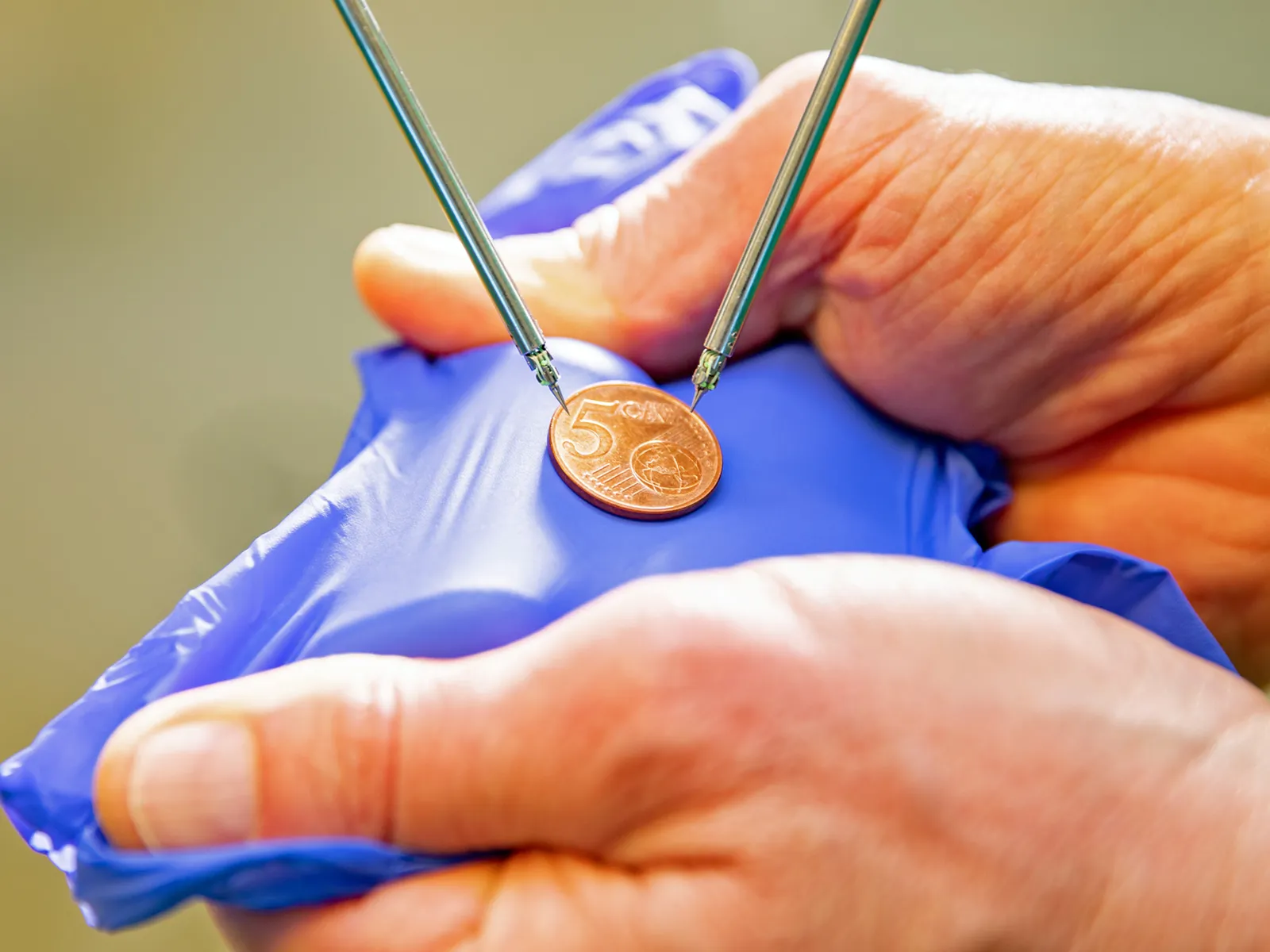 5 Cent-Münze zwischen zwei Händen auf lilafarbenem Untergrund und zwei Metallspitzen.