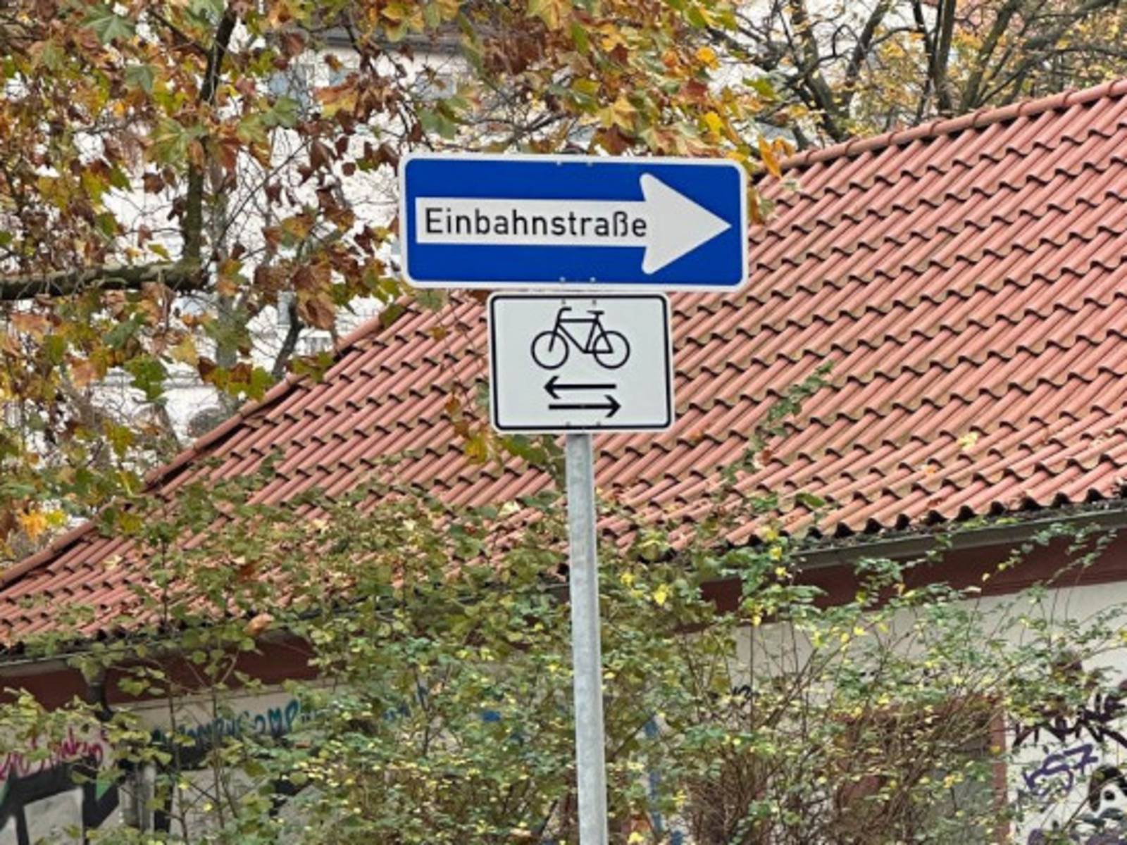 Ein blaues Schild mit einem Pfeil und dem Text Einbahnstraße, darunter ein weißes Schild mit einem Fahrradsymbol und Pfeilen nach rechts und links