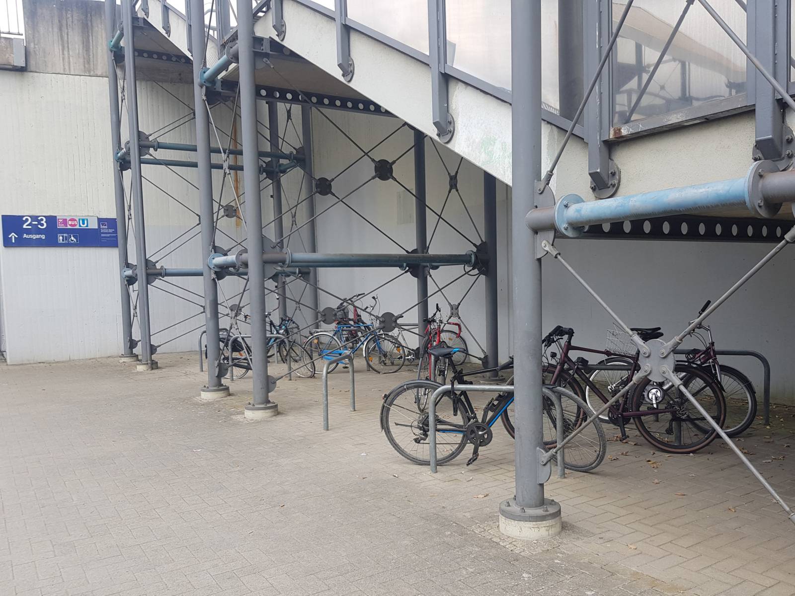 Einige Fahrradb&uuml;gel auf einem gepflasterten Platz unterhalb einer Treppe, an die B&uuml;gel sind einzelne Fahrr&auml;der angeschlossen. Hinten eine Wand mit einem blauen Schild, darauf Pictogramme wie "Bus" und "U", den Zahlen 2-3 und das Wort "Ausgang" mit einem Pfeil.