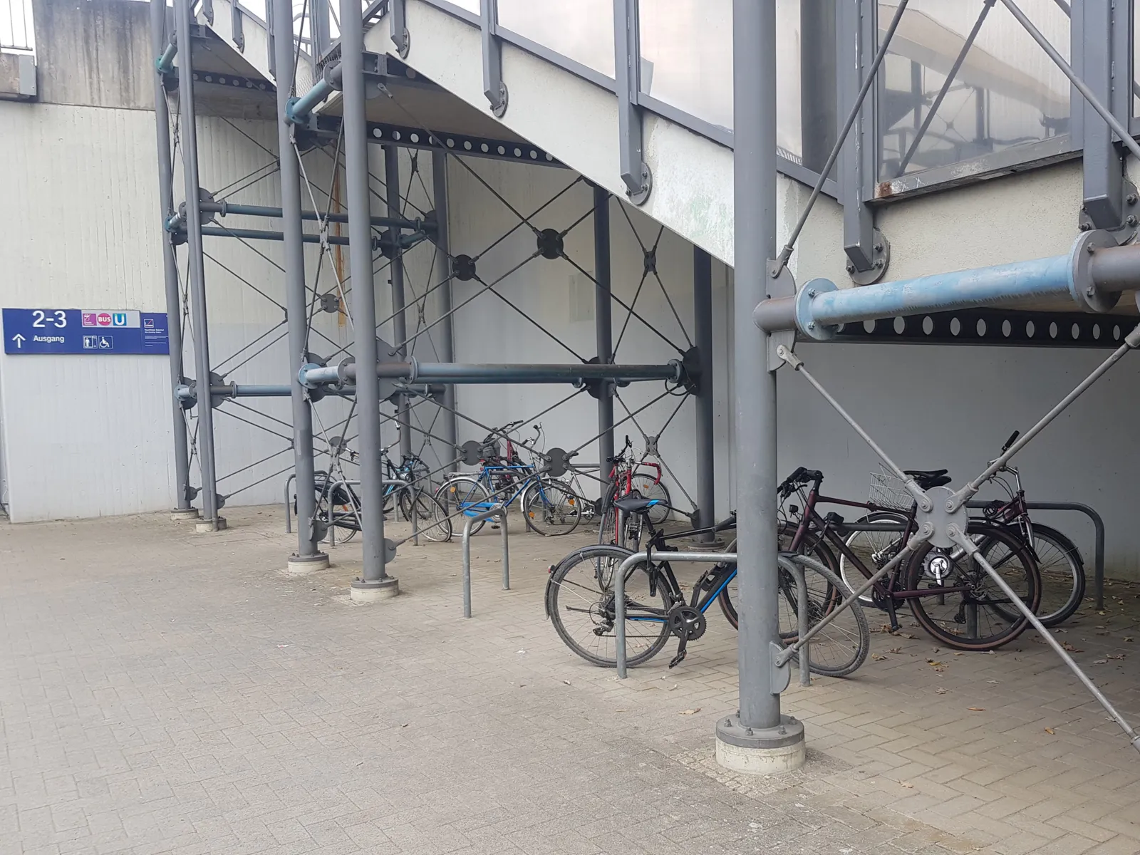 Einige Fahrradb&uuml;gel auf einem gepflasterten Platz unterhalb einer Treppe, an die B&uuml;gel sind einzelne Fahrr&auml;der angeschlossen. Hinten eine Wand mit einem blauen Schild, darauf Pictogramme wie "Bus" und "U", den Zahlen 2-3 und das Wort "Ausgang" mit einem Pfeil.