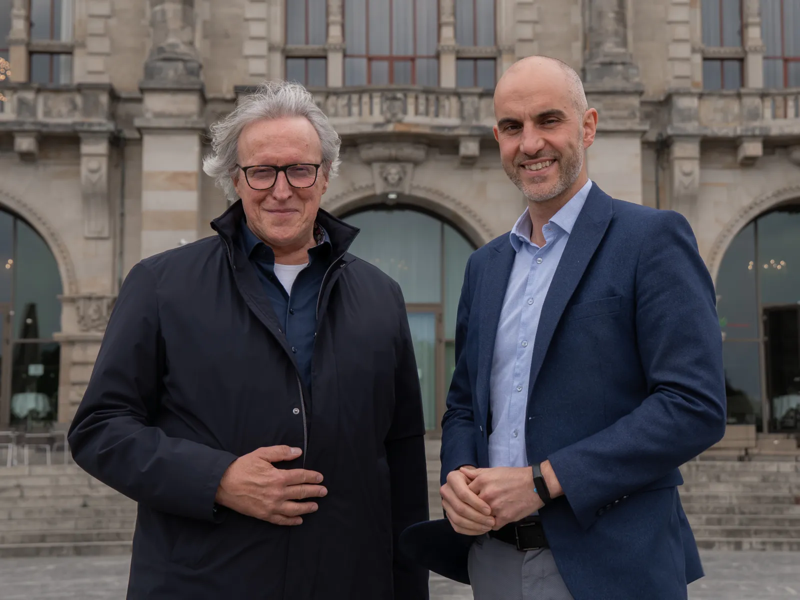 Zwei Personen schauen in die Kamera. Die linke trägt einen Windbreaker und eine Brille, die rechte einen Anzug.