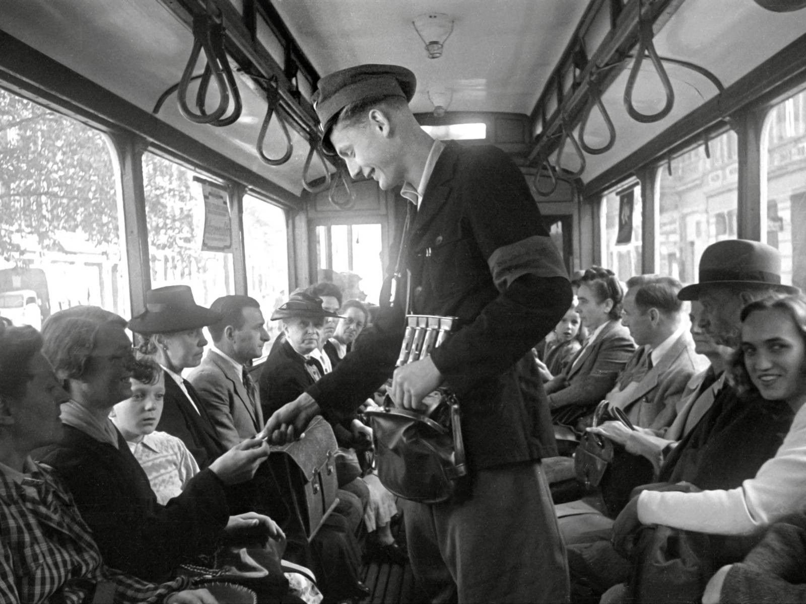 Schwarz-Weiß-Fotografie: Ein Mann trägt eine Dienstmütze und verkauft Fahrscheine gegen Bargeld in einer Straßenbahn.