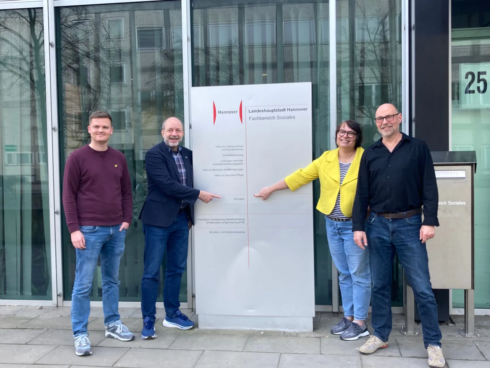 Drei Männer und eine Frau stehen vor einem Gebäude mit hohen Fenstern und einem hohen silbernen Schild mit dem Logo der Landeshauptstadt Hannover und Stellen des Fachbereichs Soziales. Ein Mann und eine Frau zeigen auf die Mitte des Schildes auf den Textzug "Wohngeld".