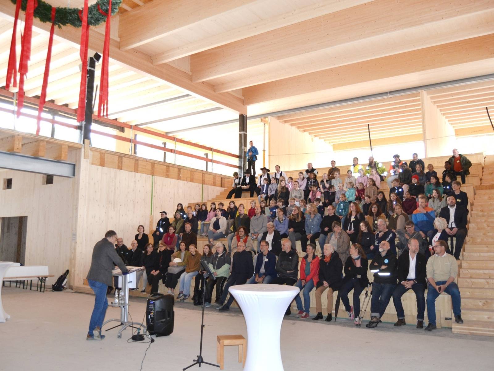 Ein gro&szlig;er Saal mit Holzw&auml;nden und Holzdecke, oben links h&auml;ngt ein Teil eines Richtkranzes. Ein Mann steht mit dem R&uuml;cken  zur Kamera gewandt vor Rednerpult, vor ihm sitzen Menschen auf mehreren Trib&uuml;nenreihen.