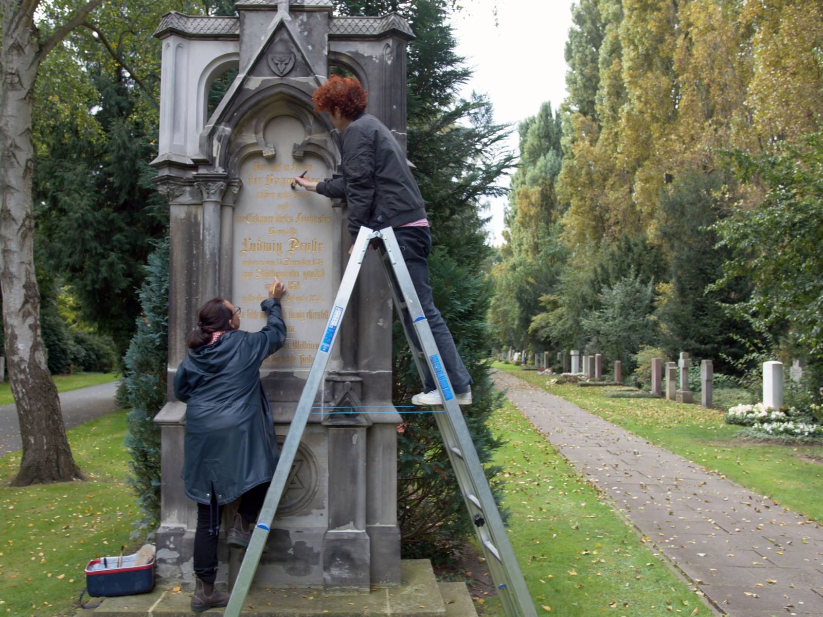 Ein Grabmahl auf dem Stadtfriedhof Engesohde, davor stehen zwei Frauen und restaurieren mit Pinseln in der Hand den Grabstein.