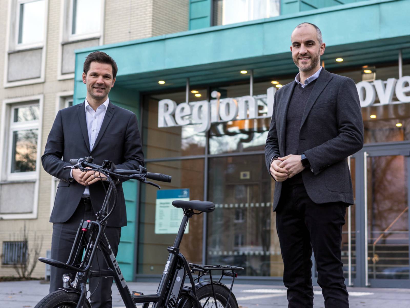 Regionspräsident Steffen Krach (links) und Oberbürgermeister Belit Onay (rechts) stehen vor dem Regionshaus, zwischen ihnen steht ein Elektrisches, zusammenfaltbares Fahrrad.