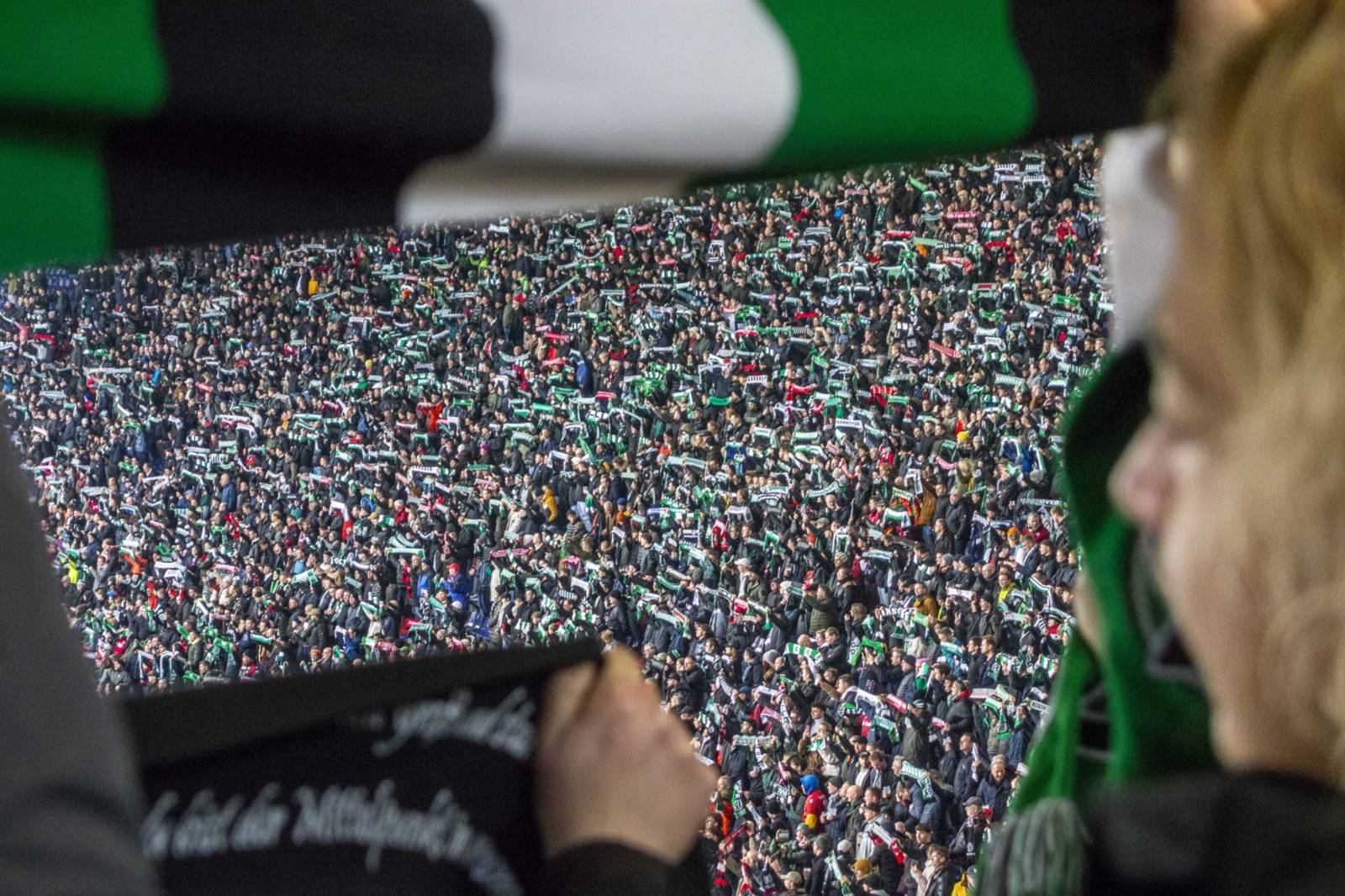Zu sehen ist eine Fotografie eines vollen Fu&szlig;ballstadions, am rechten Rand erkennt man eine Person, die in die Menge hineinschaut.