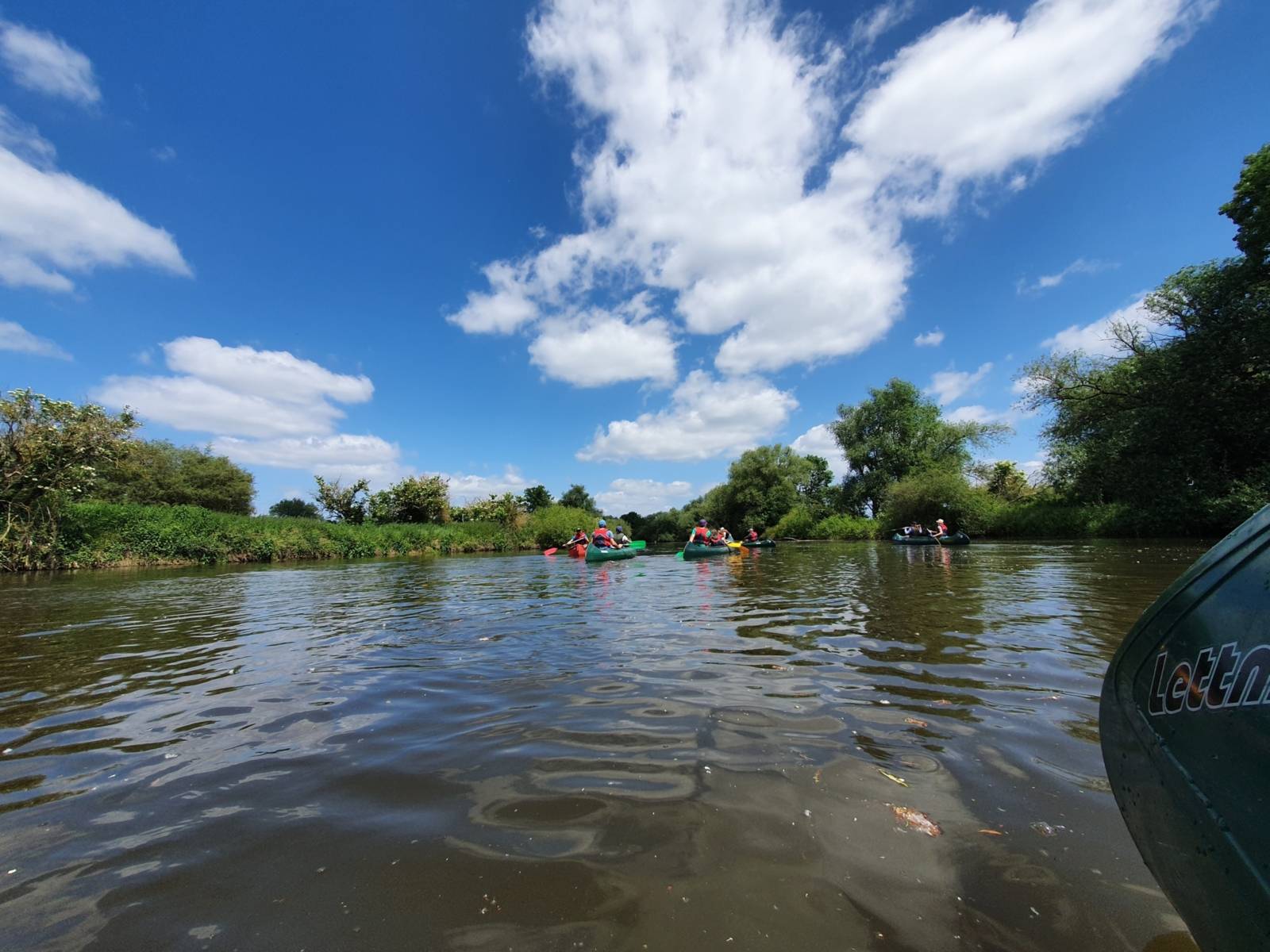 Blick nach vorn aus der Mitte des Flusses: In der Entfernung sechs bunte Paddelboote, rechts und links die grünbewachsenen Ufer, ein blauer Himmel mit Zirruswolken