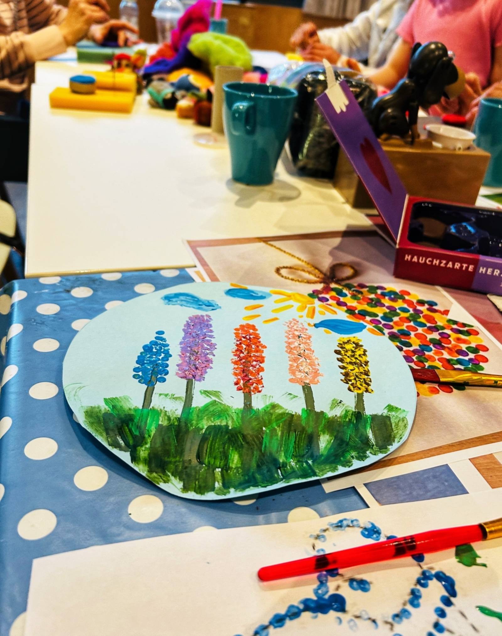 Ein Tisch mit Bastelsachen im Hintergrund. Im Vordergrund Ist ein blauer Kreis aus Pappe, darauf sind Blumen gemalt.
