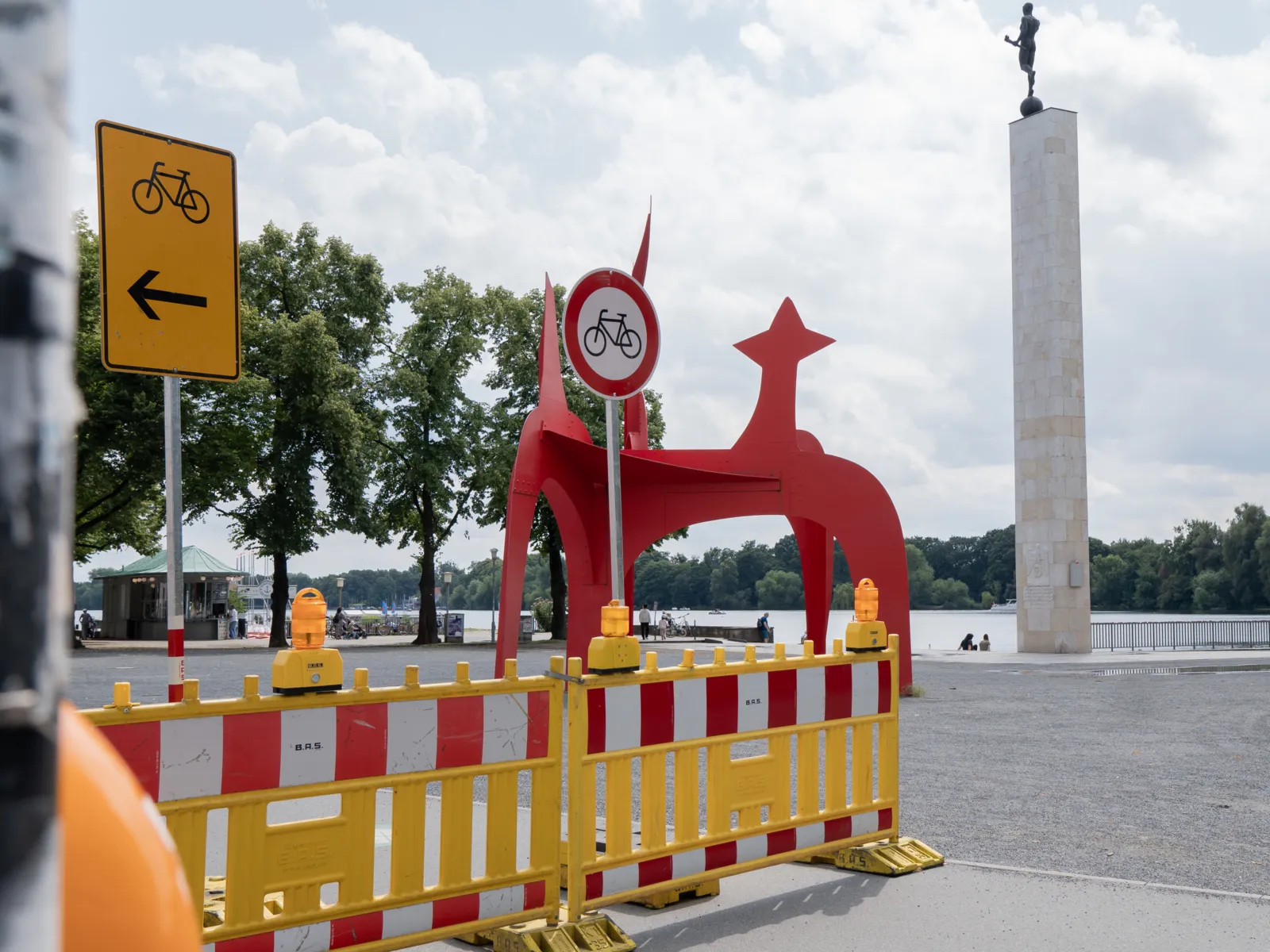 Eine gelbe aufgestellt Absperrung mit rot-weißen Streifen auf einem Weg. Darüber ein Verkehrsschild Radfahren verboten. Im Hintergrund eine rote, große Kunstskulptur, eine hohe Säule mit einem Staffelläufer, dahinter ein See.
