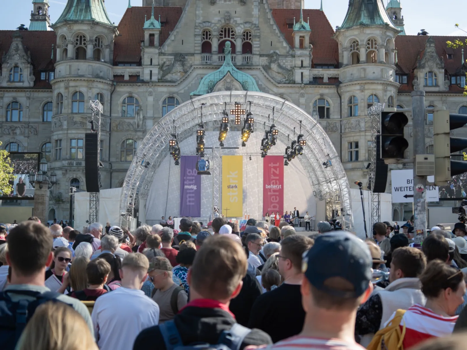 Viele Menschen stehen vor einer Bühne, auf der Banner mit den Worten mutig, stark, beherzt herabhängen. Dahinter ein Gebäude.
