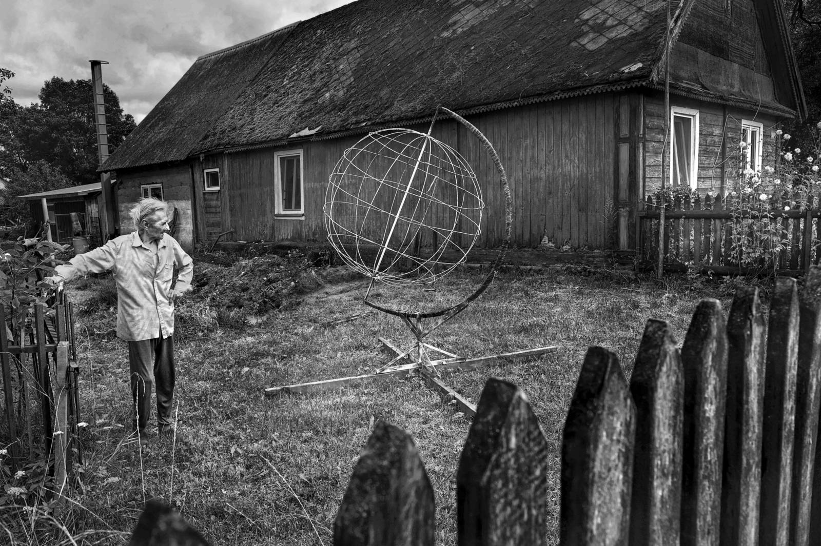 Zu sehen ist eine Fotografie von Tomasz Tomaszewski: ein Mann steht im Garten eines Hauses, ein Metallger&uuml;st, das an einen Globus erinnert, ist neben ihm zu sehen.