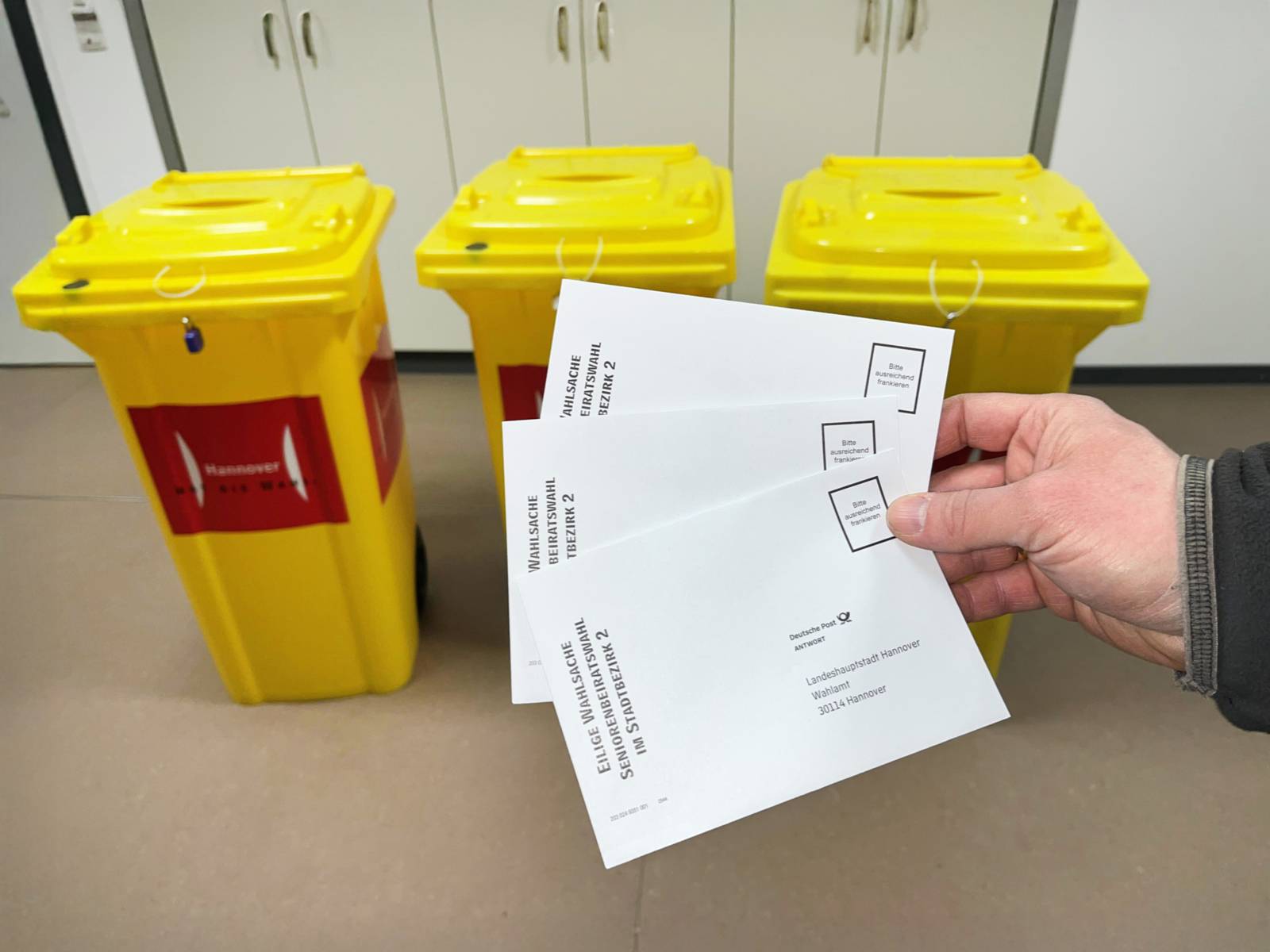 Eine Hand hält drei Briefumschläge, dahinter stehen drei gelbe Container oben mit offenen Einwurfschlitzen, vorn ein roter Aufkleber mit dem Logo Landeshauptstadt Hannover