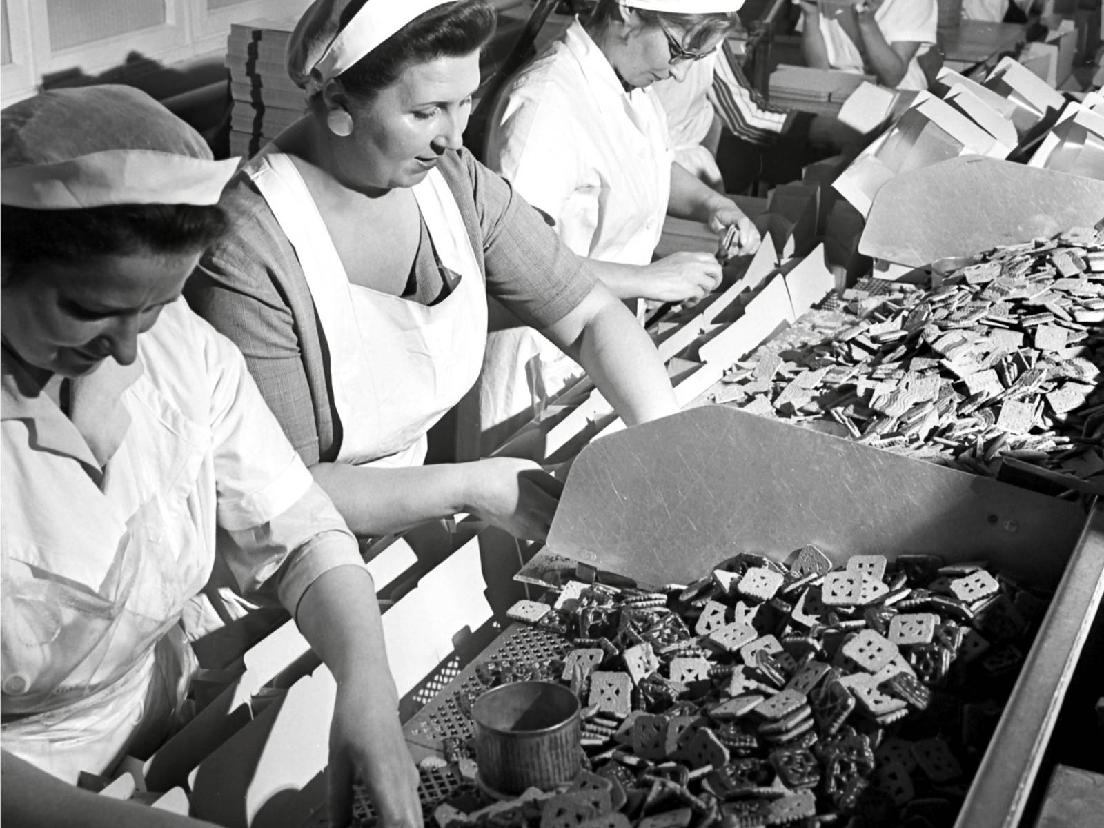 Schwarz-Weiß-Fotografie: Frauen sitzen nebeneinander und sortieren Kekse.