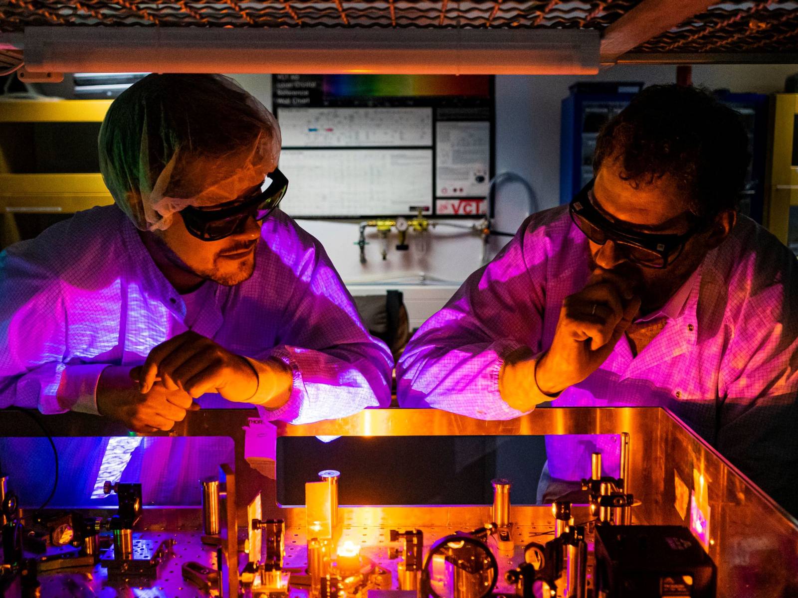 Zwei Männer mit Schutzbrillen in orange-lilalem Licht vor einem optischen Experiment.