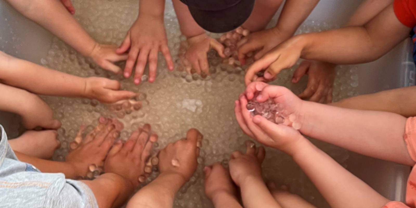 Hände von zehn Kindern greifen schwimmende Kügelchen in einer Wanne mit Wasser.