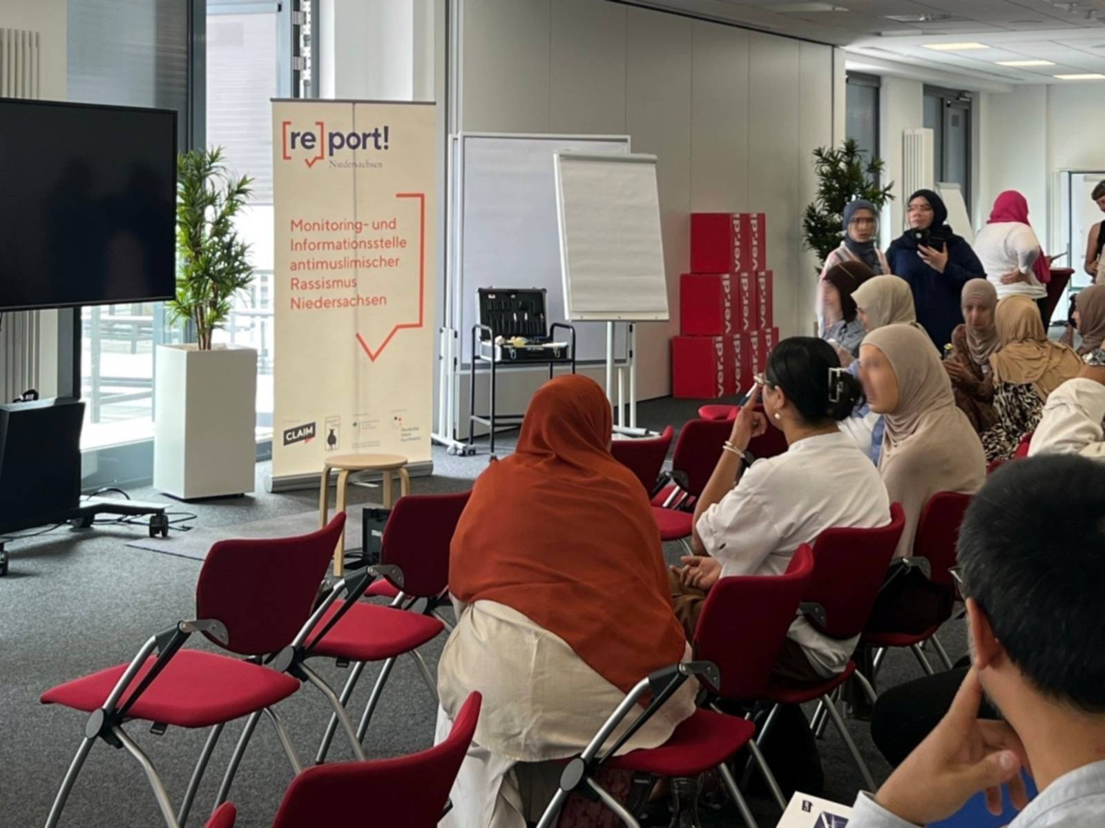 Ein heller Konferenzraum, in dem einige Menschen auf Stuhlreihen sitzen. Die Menschen sind von schräg hinten fotografiert. Es handelt sich anscheinend größtenteils um Frauen. Vor den Stuhlreihen ein Flip-Chart mit dem Text "report Niedersachsen. Monitoring- und Informationsstelle antimuslimischer Rassismus Niedersachsen".