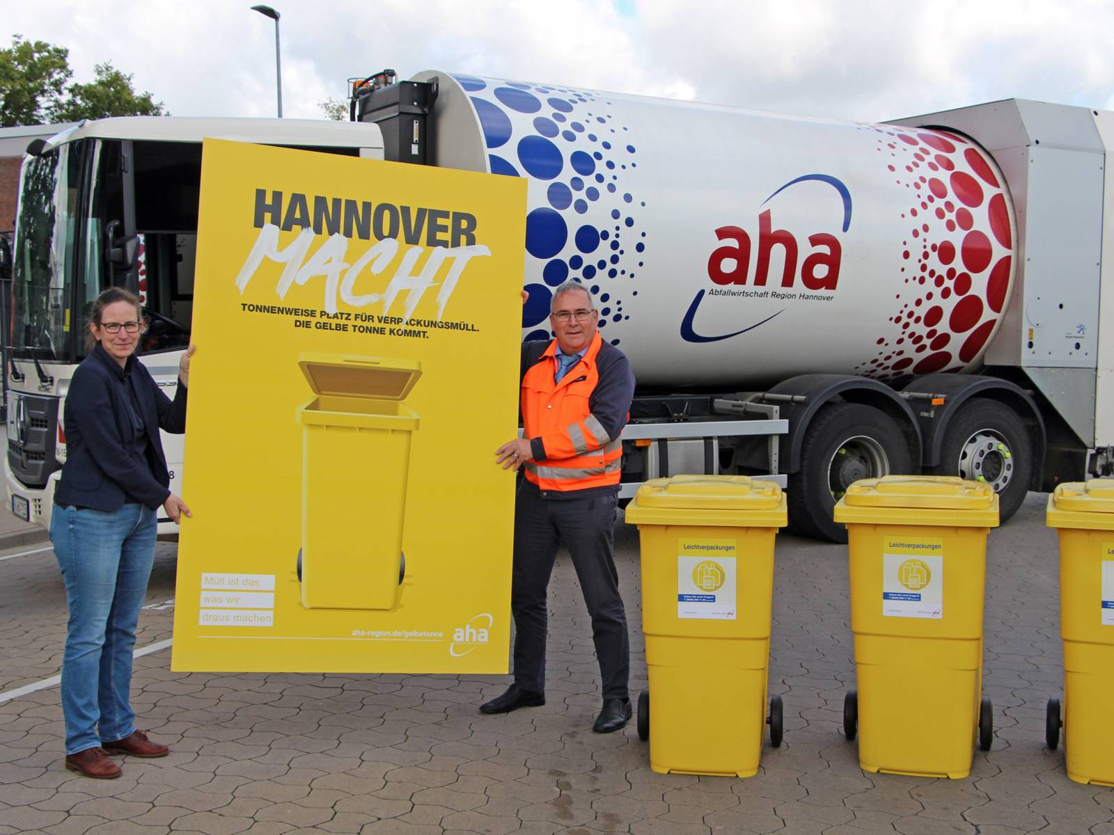 Eine Frau und ein Mann vor einem Müllauto stehend halten ein großes gelbes Plakat mit der Aufschrift "Hannover macht" in den Händen, daneben stehen drei gelbe Mülltonnen mit der Aufschrift "Leichtverpackungen".