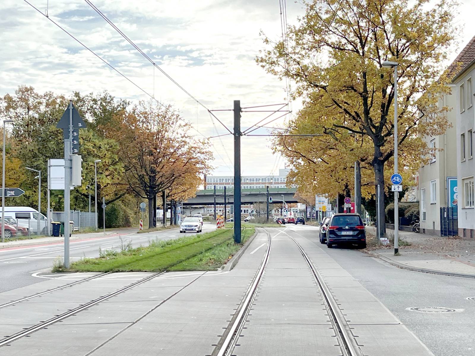 Eine Straße mit einem Mittelstreifen mit grünem Gras, in der Mitte verlaufen Schienen, oben Stromleitungen. Rechts parken Autos.