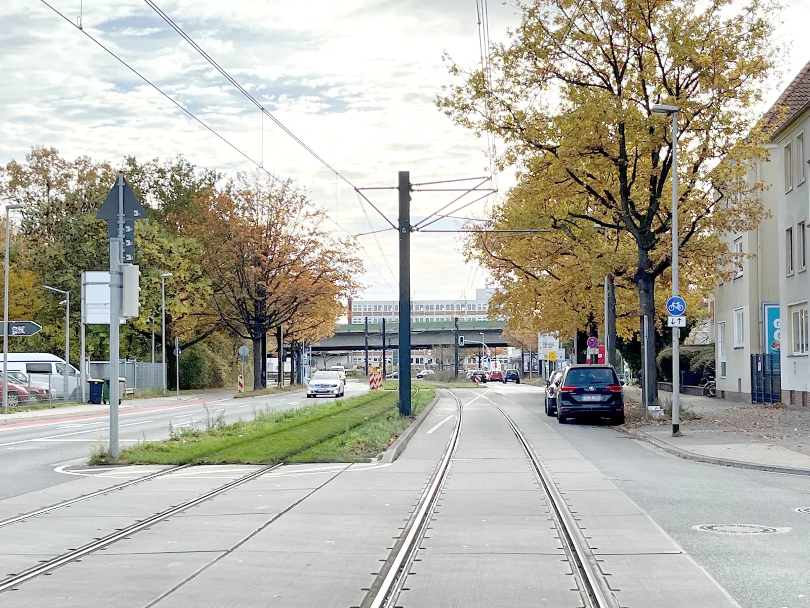 Eine Straße mit einem Mittelstreifen mit grünem Gras, in der Mitte verlaufen Schienen, oben Stromleitungen. Rechts parken Autos.