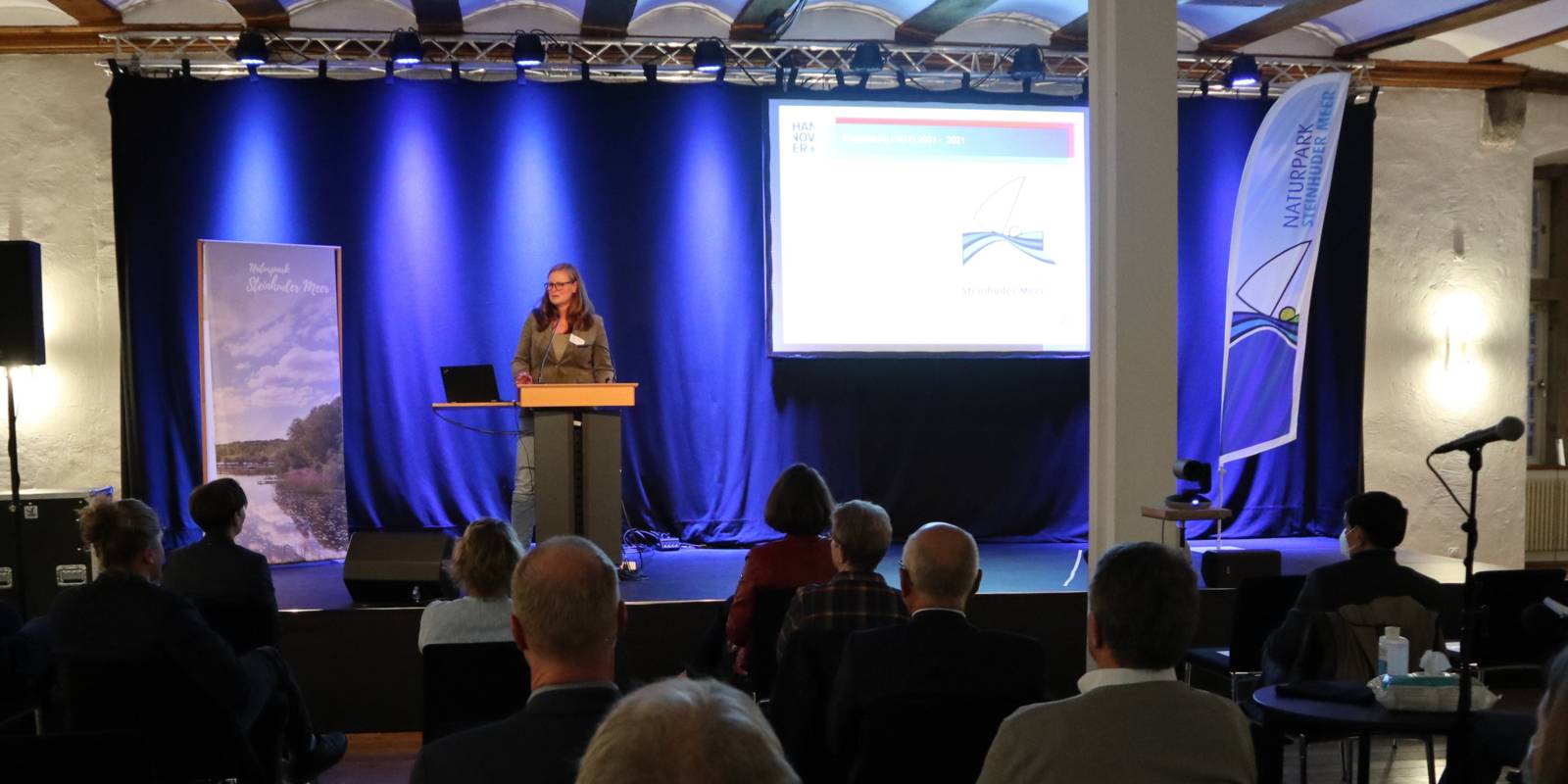 Eine Frau steht auf einer Bühne hinter einem Redepult. Publikum sitzt davor.