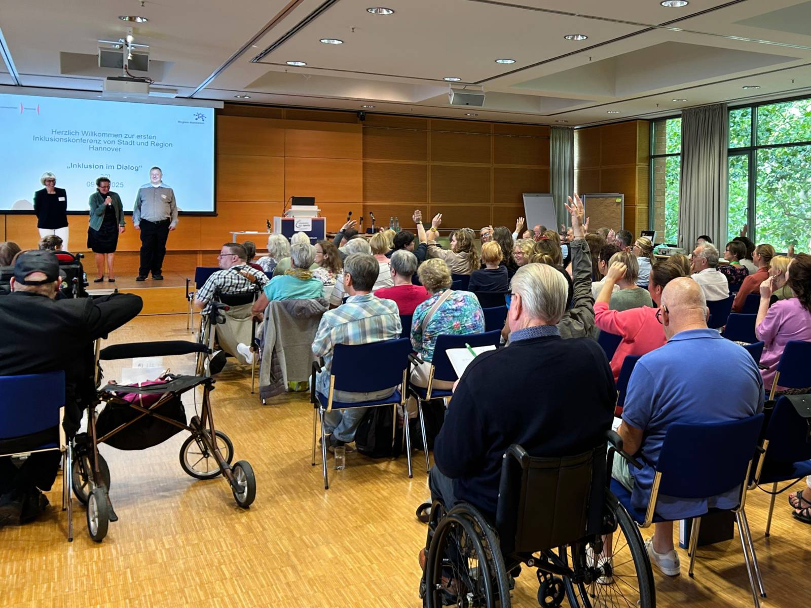 Drei Personen stehen auf einem Podest vor einem Publikum, das von hinten zu sehen ist und auf Stühlen und einem Rollstuhl sitzt. Einige Personen heben die Hand.